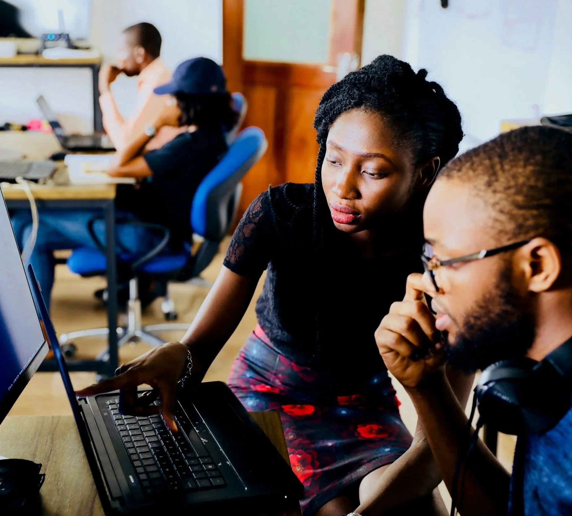 Two young adults working together on a laptop in an office, with three other people working at desks in the background.