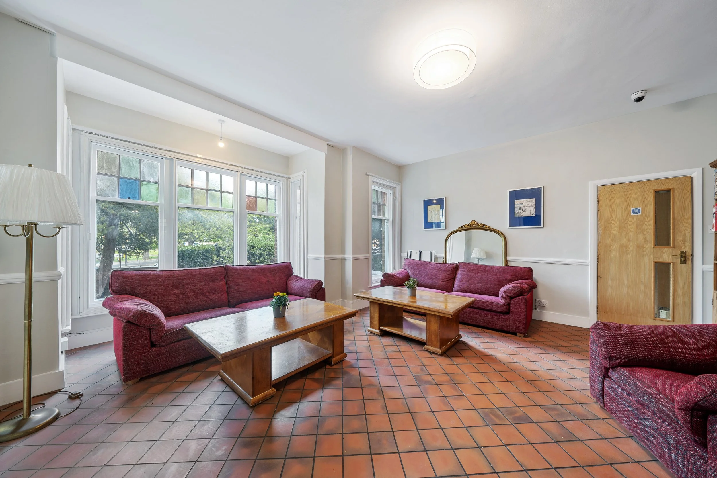 Lounge area with three red sofas, wooden coffee tables, a large bay window with stained glass, and artwork on the walls.
