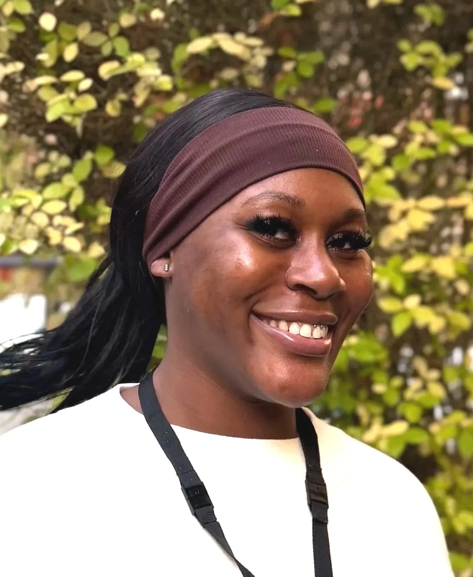 A woman smiling outdoors with a blurred background of green and yellow leaves.