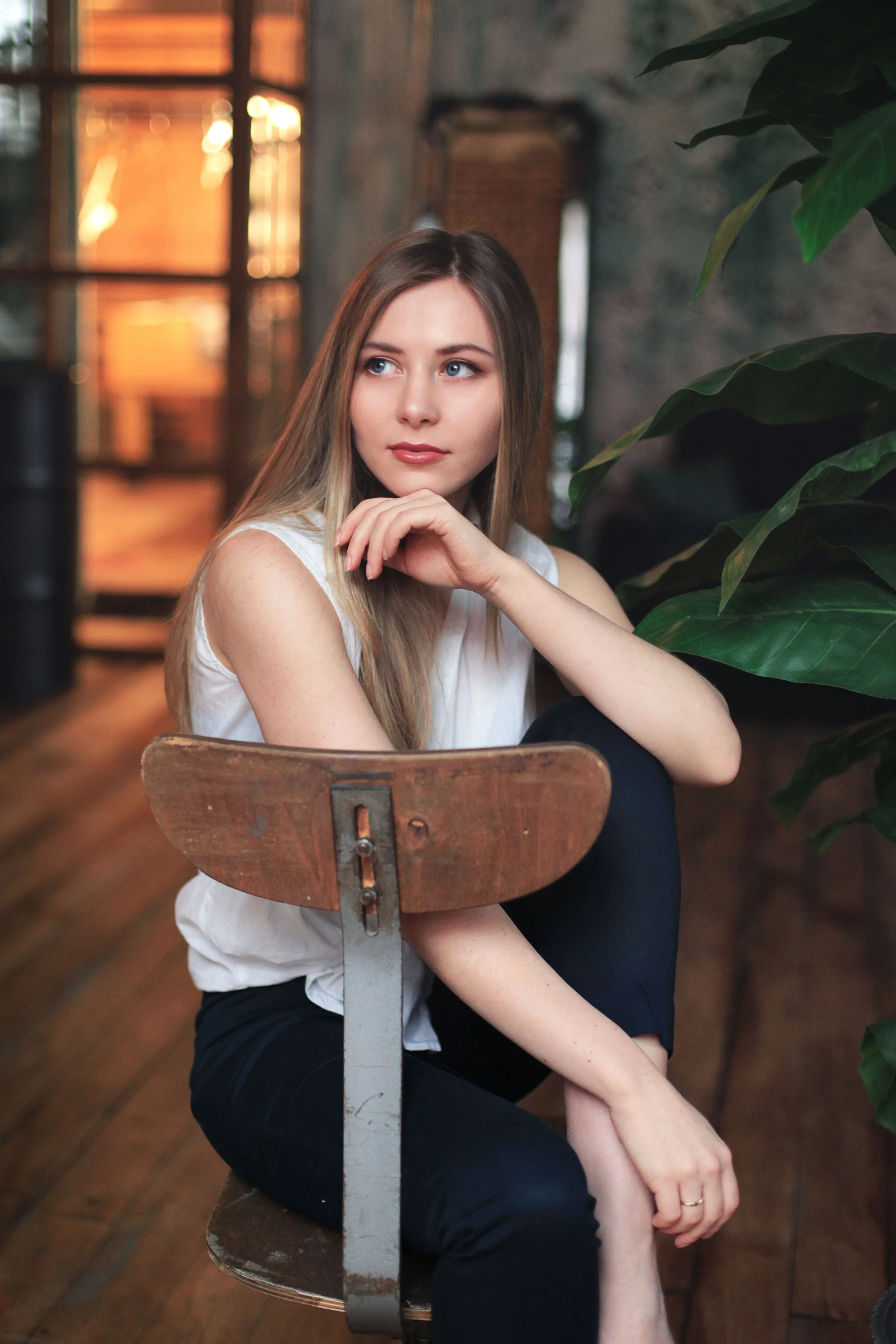 A young woman with long blonde hair, sitting on a wooden chair with a metal backrest, in a cozy, dimly lit indoor space with brick walls and lush greenery.