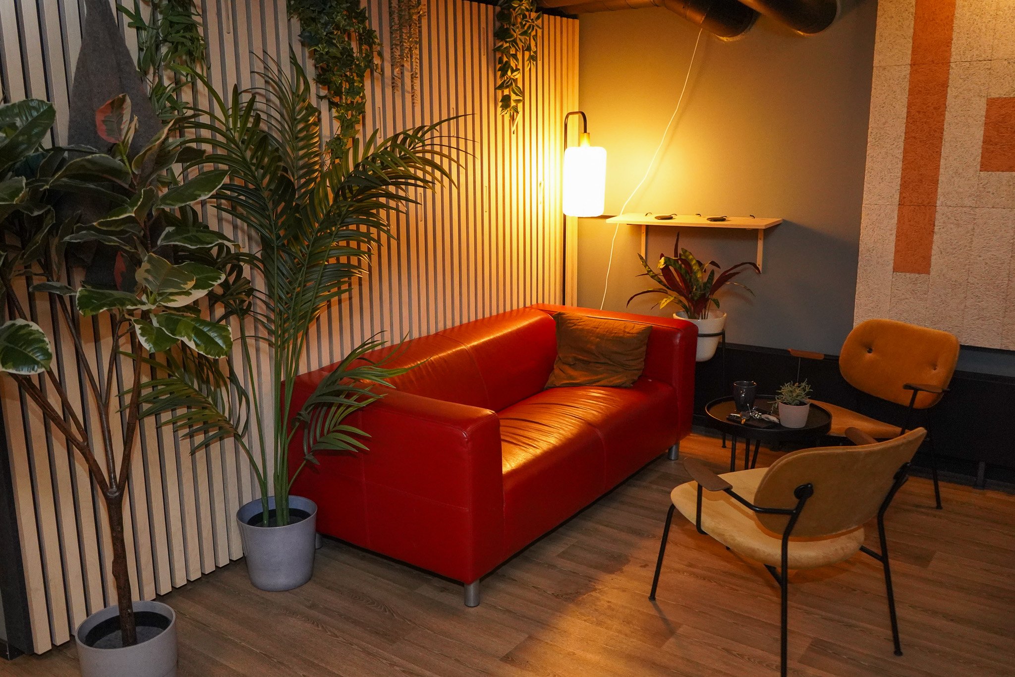 Innendørs stue med rød sofa, to beige stoler, planter, gul belysning og trehjul på gulvet.