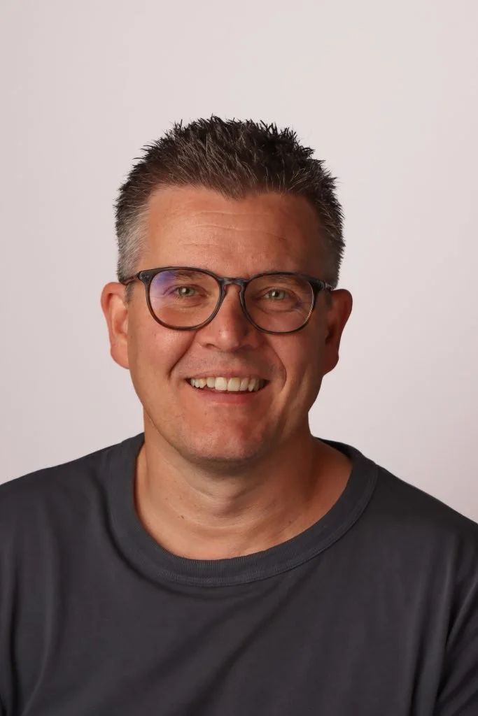 Smilende mann med kort, grått hår og briller, iført svart t-skjorte, mot en lys bakgrunn.