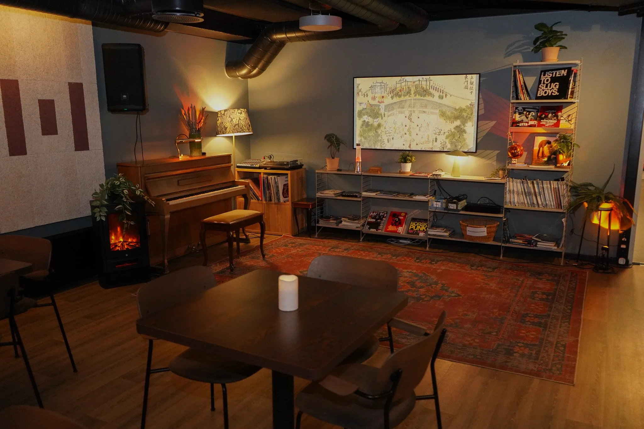 Et koselig rom med et piano, en vedovn, en sofa, en stuepult med bøker og filmer, og et rundt bord med lys på. Det er dekorative planter, en gul lampe, og en vegg med kunstverk. Rommet har tregulv og en stor rød teppe.