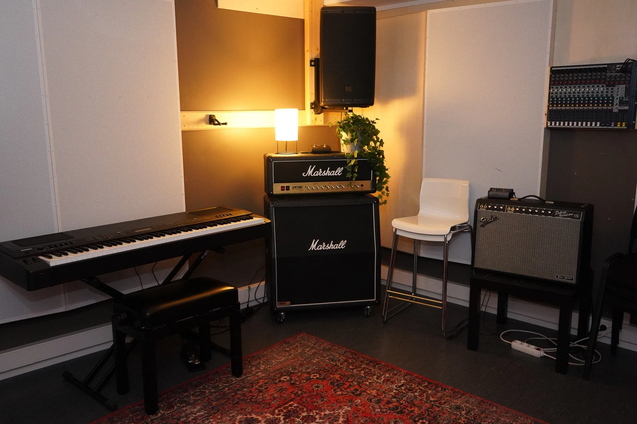 Innendørs musikkrom med keyboard, to Marshall forsterkere, en hvit stol, en liten stumtjener, og en rød teppe på gulvet.