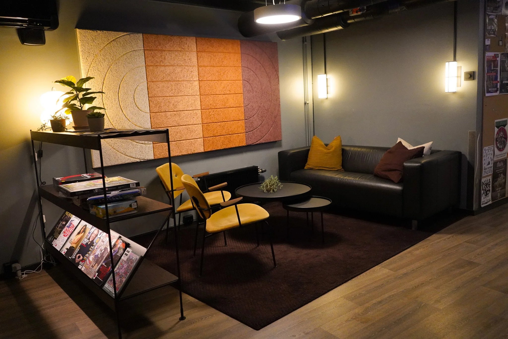 En komfortabel lounge med svart sofa, fargede puter, gule stoler, et lite svart bord med dekorasjon, en svart metallhylle med magasiner, og et stort abstrakt kunstverk på veggen.