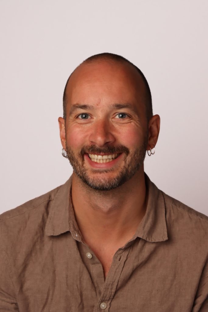 Porträtt av en man med skägg, piercings i öronen, kort hår och brunt skjorta, som ler mot kameran.