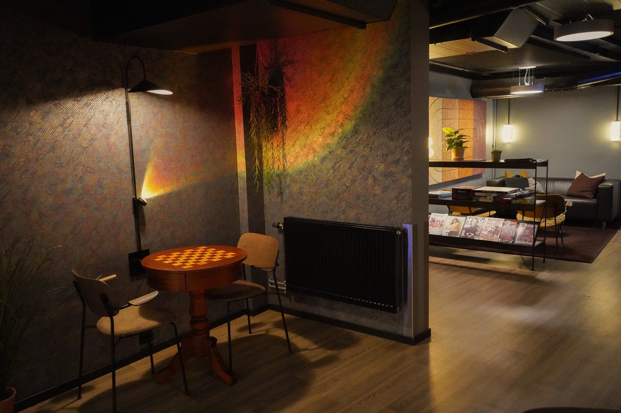 Et koselig rom med et lite bord og tre stoler, et vegghengt radiator, en vegg med fargerik tapet og en plante. I bakgrunnen er det en annen sittestue med sofa, lerreter med bøker, og lysende vegglamper i et moderne interiør.