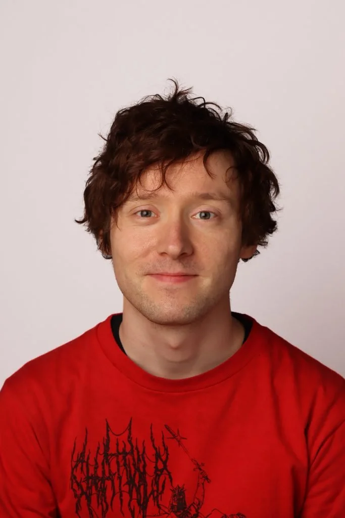 Portrett av en mann med brunt, krøllete hår, iført en rød t-skjorte med svart grafikk, mot en lys bakgrunn.