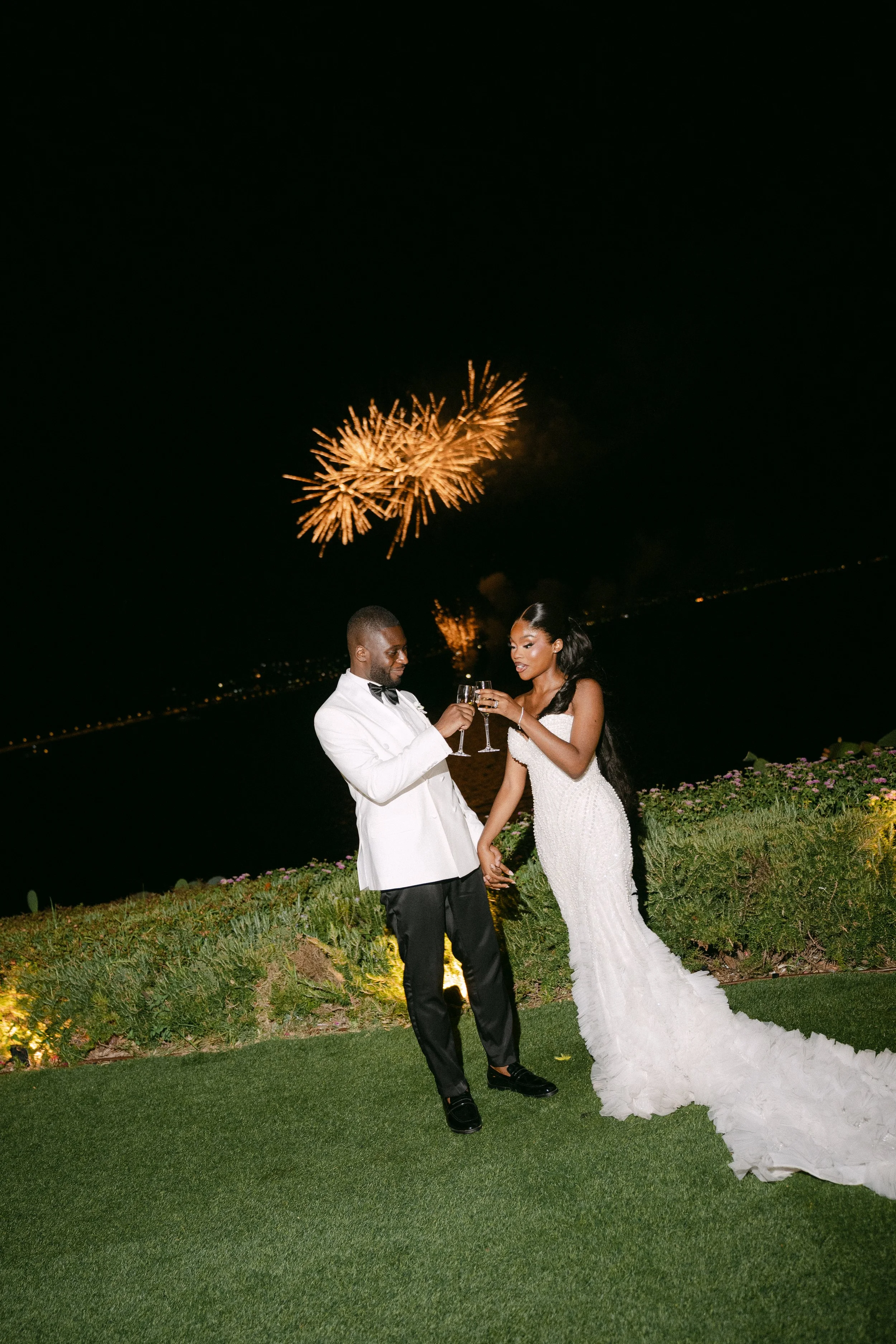 A newlywed couple toasting with champagne glasses at night with fireworks in the sky behind them, outdoors on a grassy area.