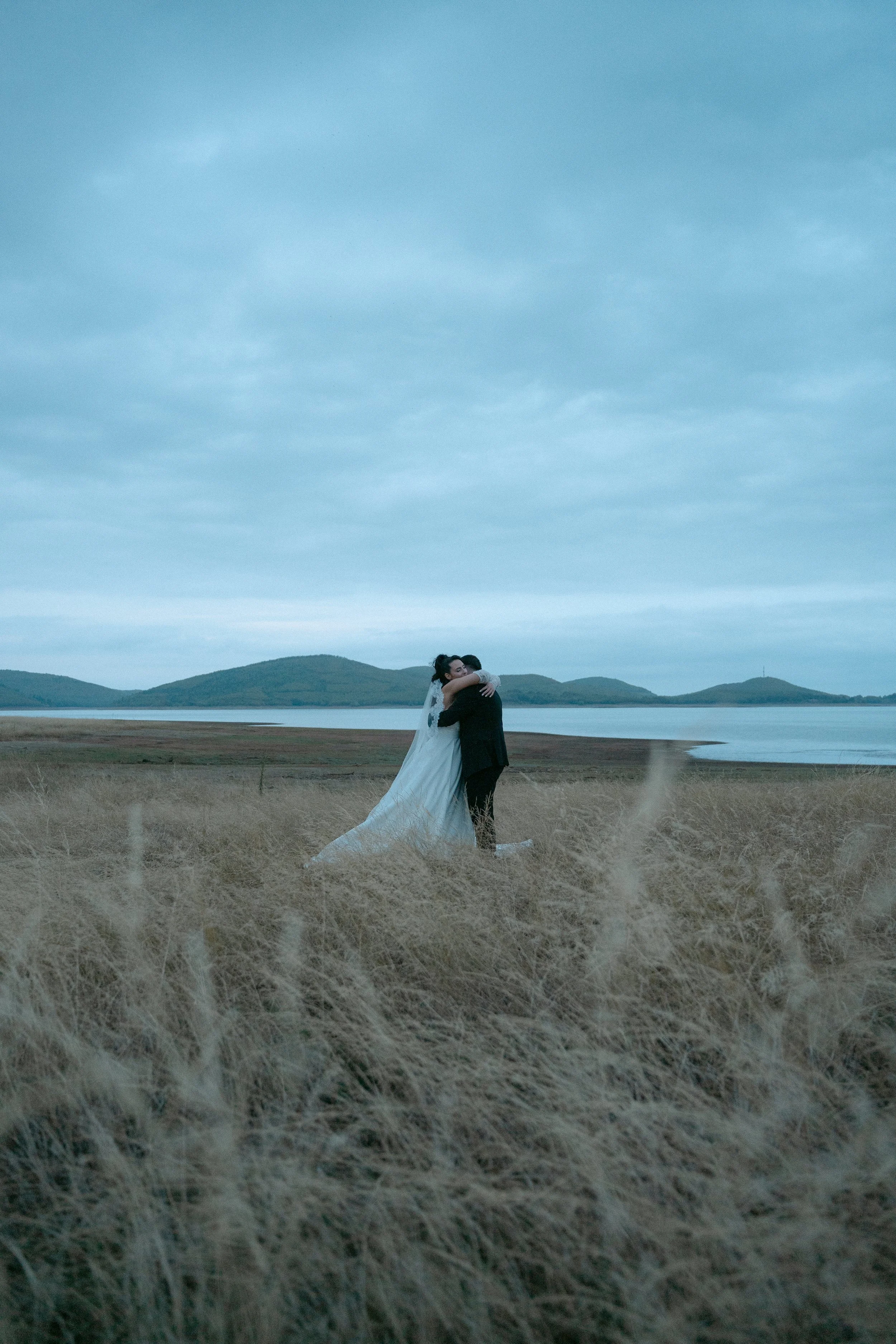 A bride and groom hugging on a grassy field near a body of water with mountains in the background