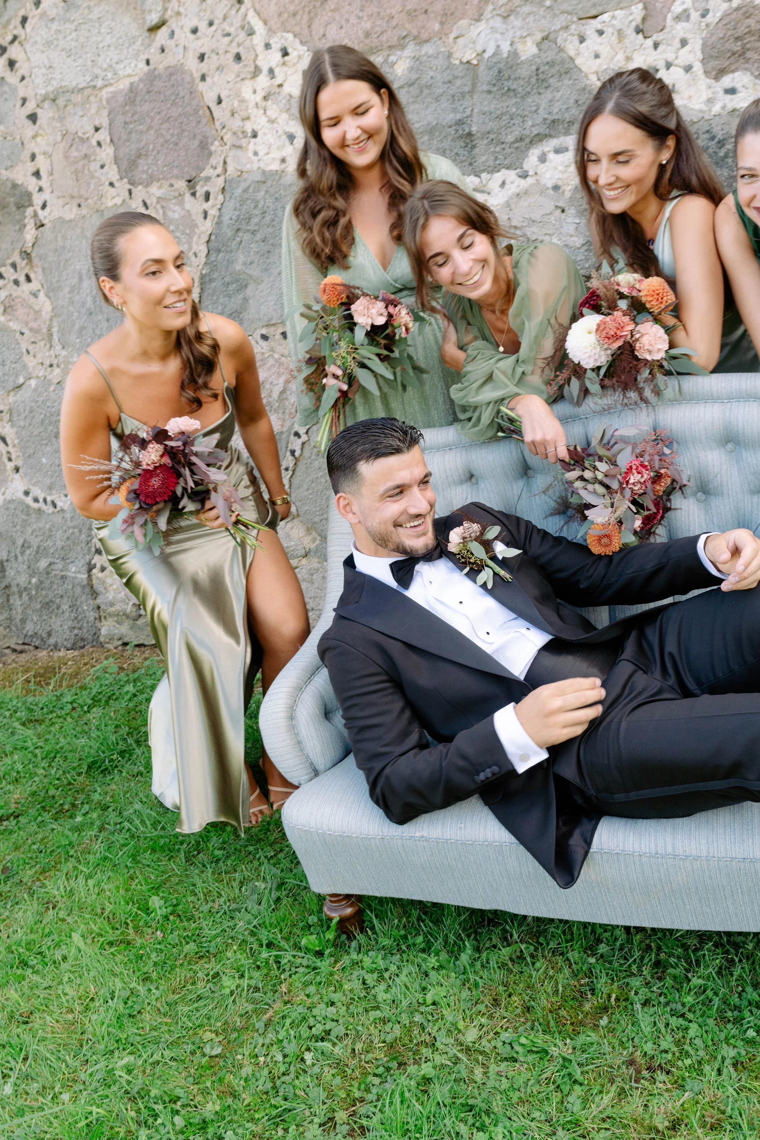 Group of women in dresses with bouquets surrounding a man in a tuxedo relaxing on a sofa outdoors.