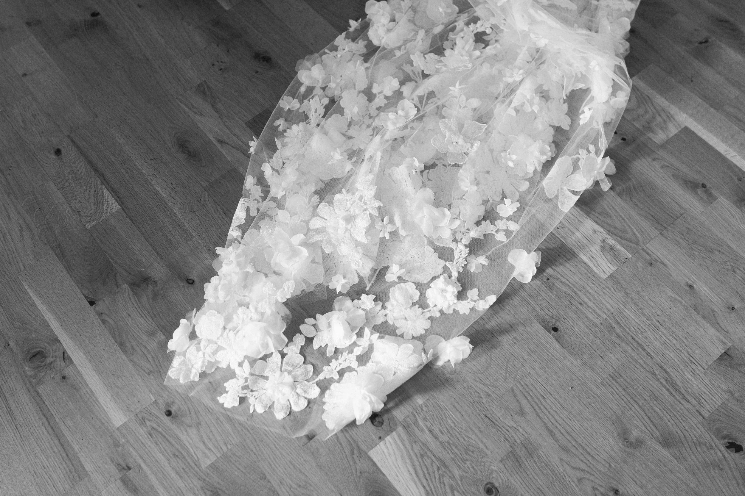 A delicate wedding veil with floral lace appliqué lies on a wooden floor.
