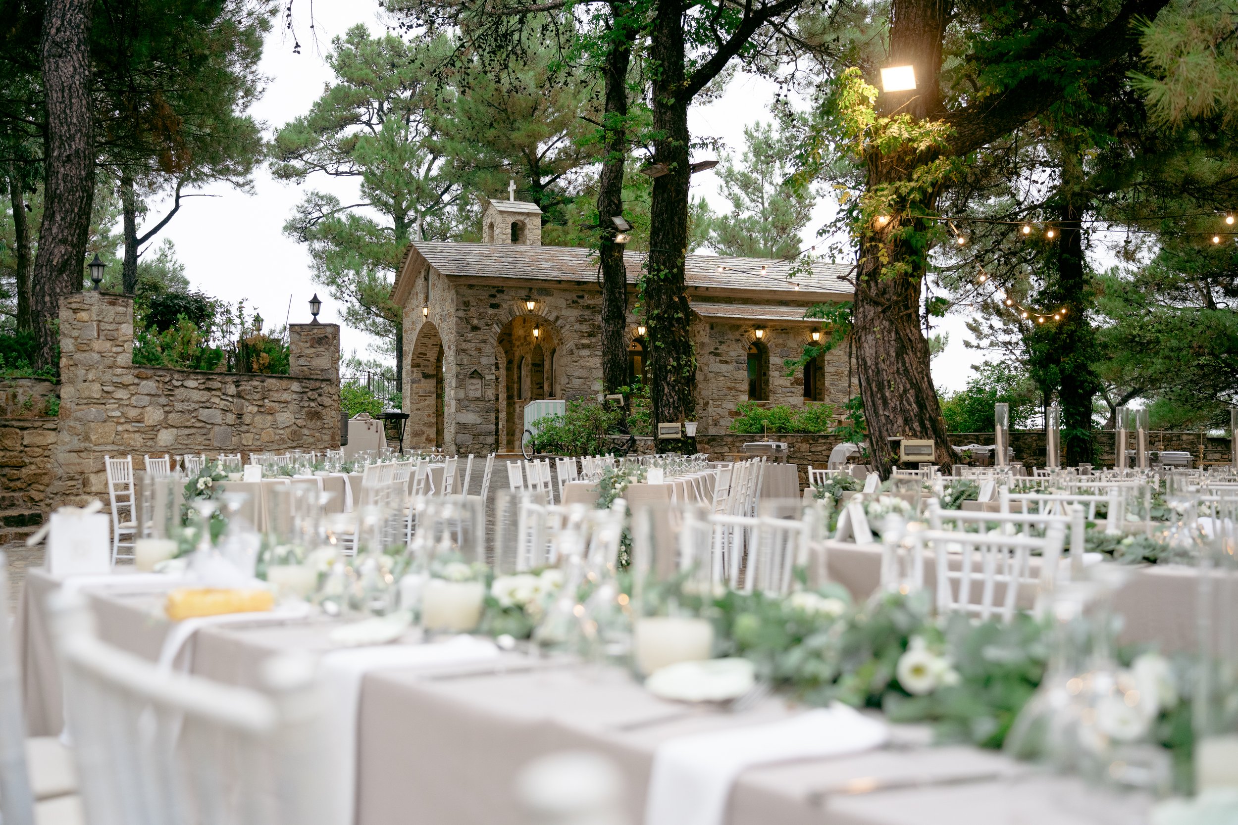 Outdoor wedding reception setup with white chairs and tables decorated with floral centerpieces, set in a garden with large trees, string lights, and a small stone chapel in the background.