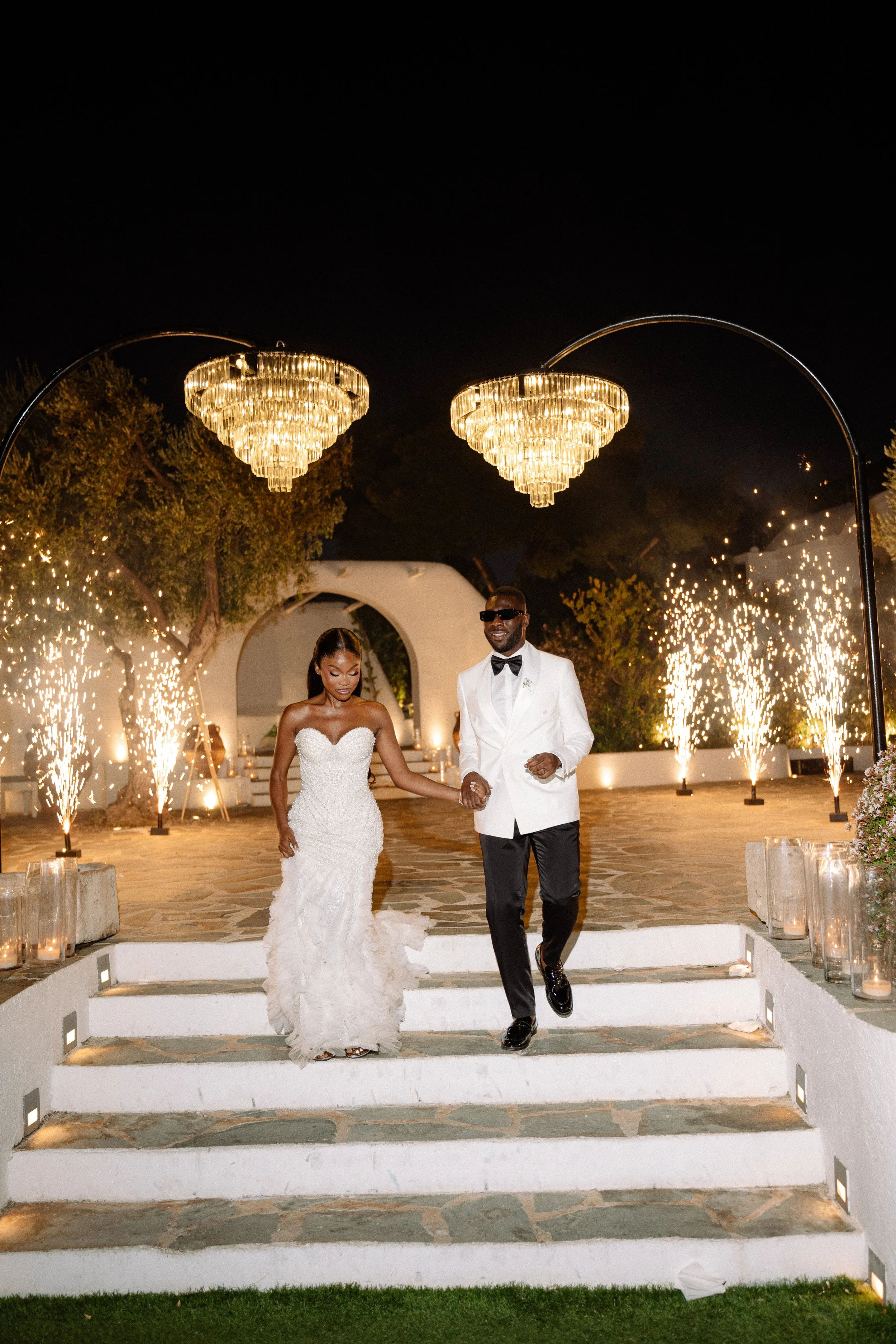 Couple in wedding attire walking down the steps of an outdoor wedding venue at night, with fireworks and chandeliers in the background.