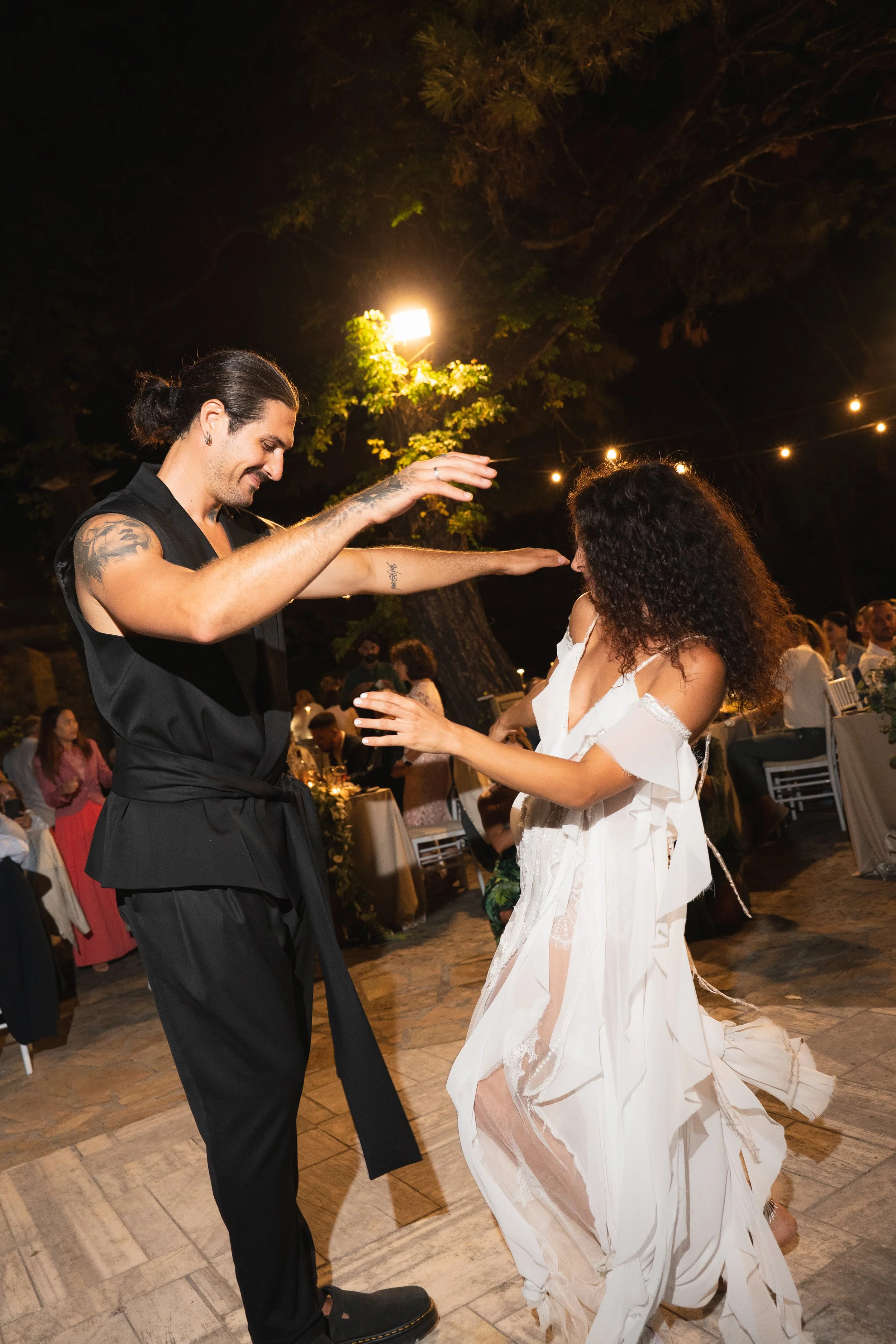 A man and a woman are dancing together at an outdoor evening event, with string lights and a tree in the background, and guests seated at tables.