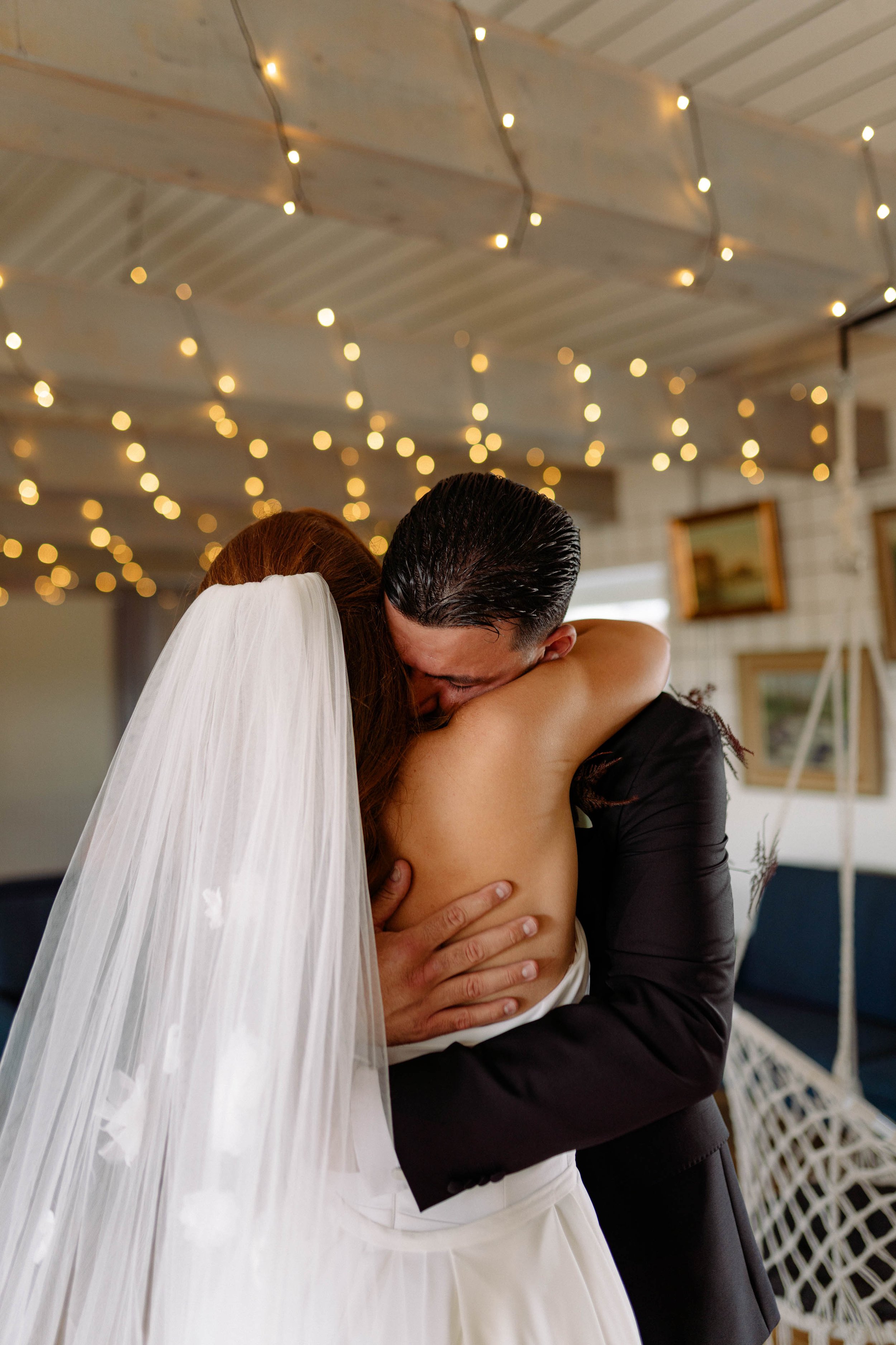 A bride and groom embrace in a heartfelt hug at their wedding, with the bride wearing a veil and the groom in a black suit, surrounded by warm fairy lights and artwork in the background.