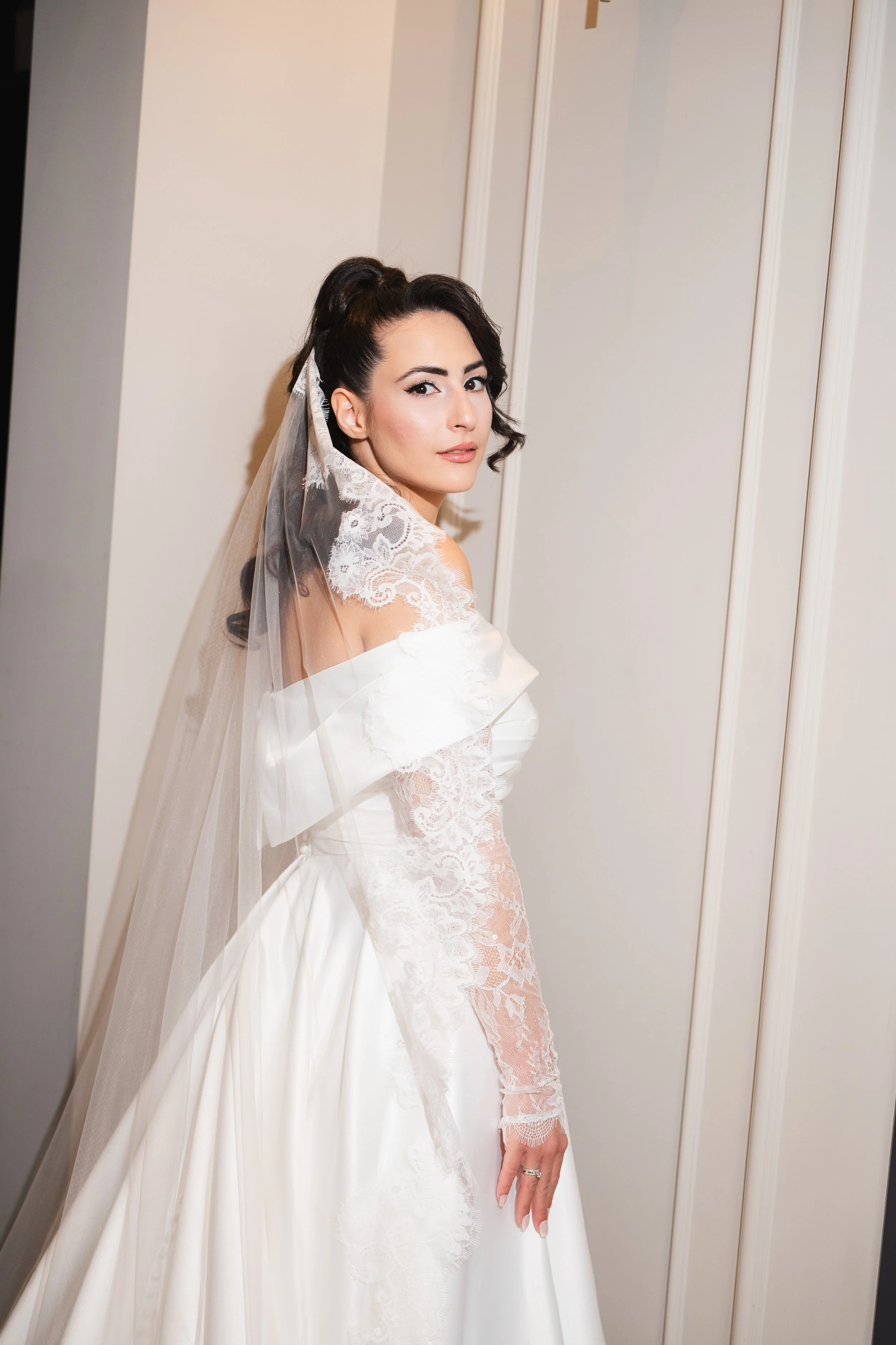 A bride in a white wedding dress with lace sleeves and a veil, standing against a plain wall, looking at the camera with a confident expression.