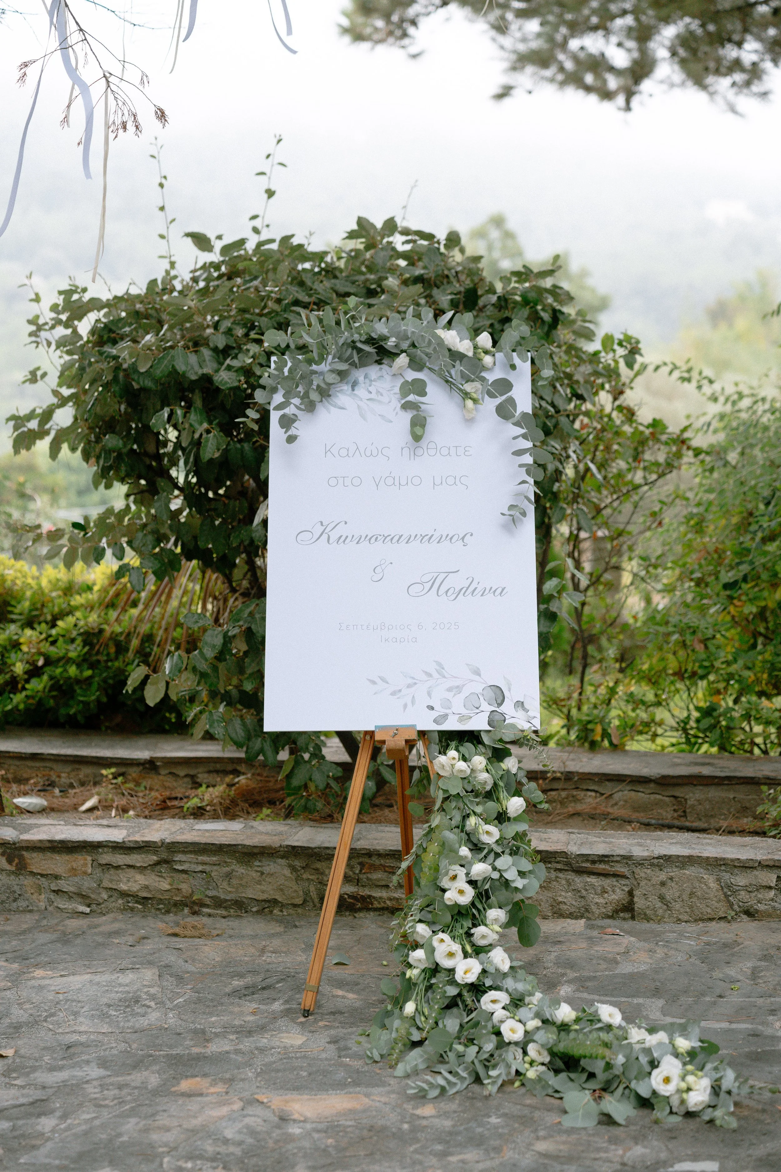 A wedding sign decorated with white flowers and greenery on an easel outside, with a stone pathway and lush garden in the background.