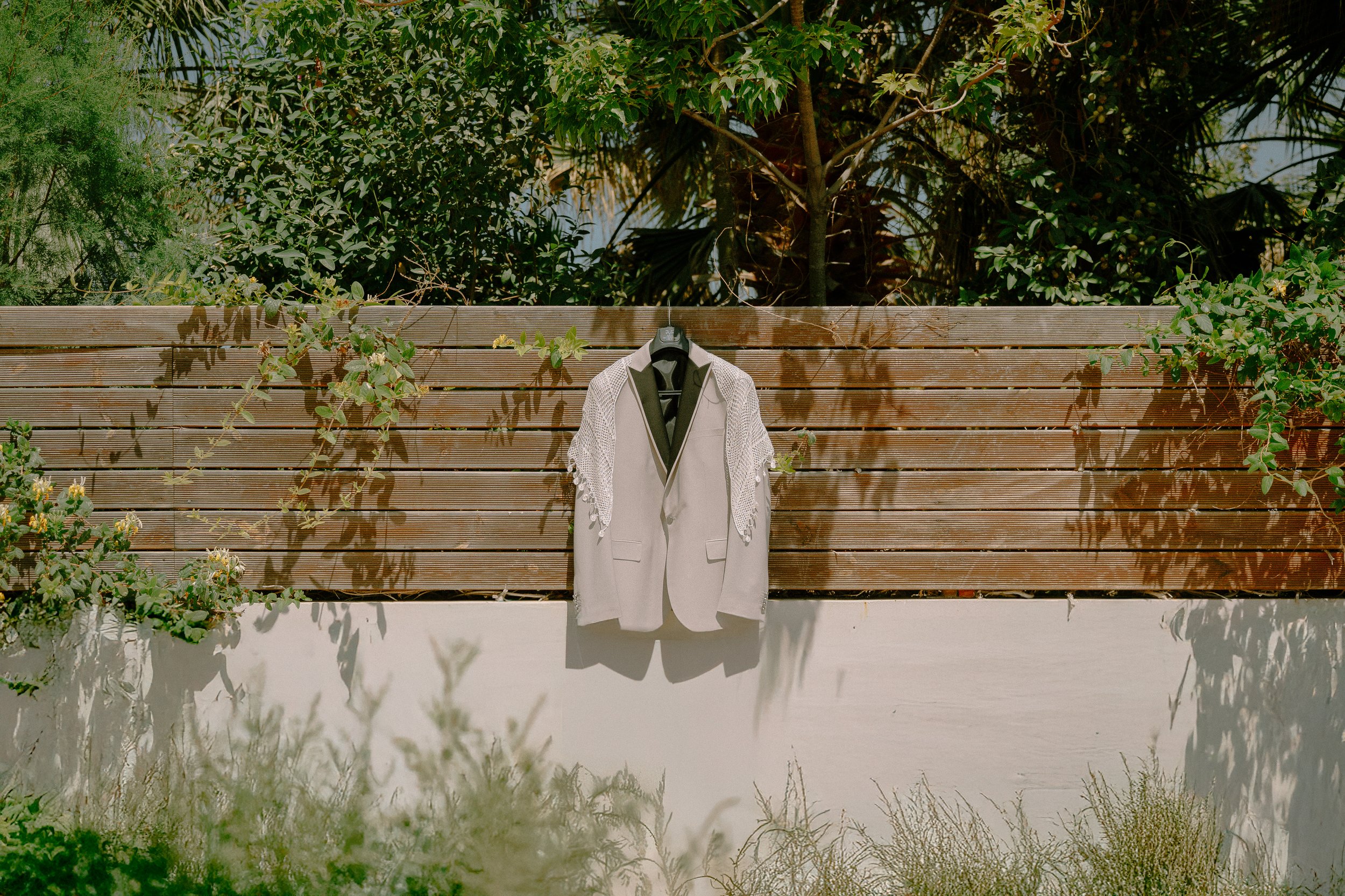 A beige blazer hanging on a black hanger against a wooden fence with greenery and plants in the background.