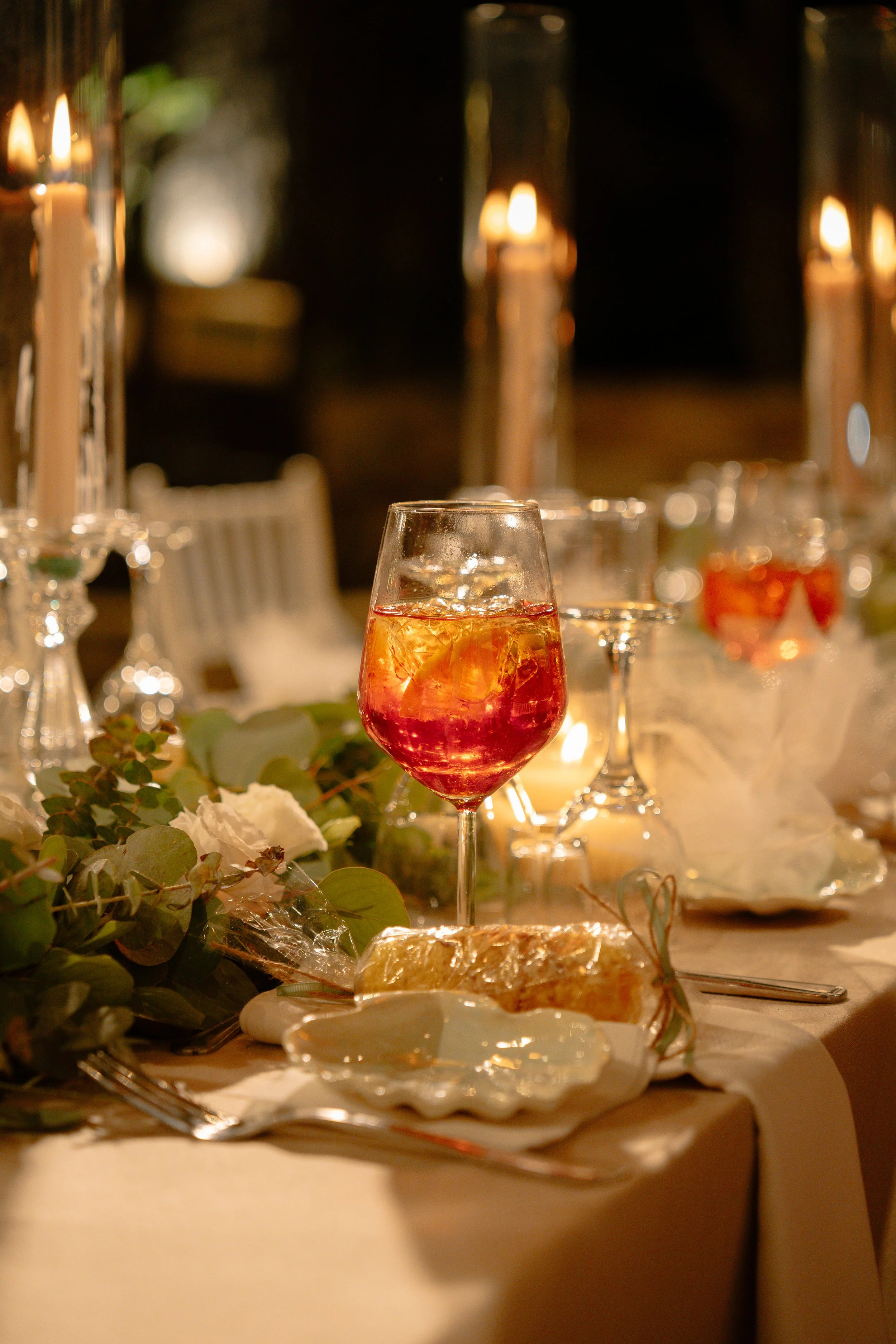 Elegant dinner table setting with a glass of red wine and ice, surrounded by candles, greenery, and a bun on a plate.