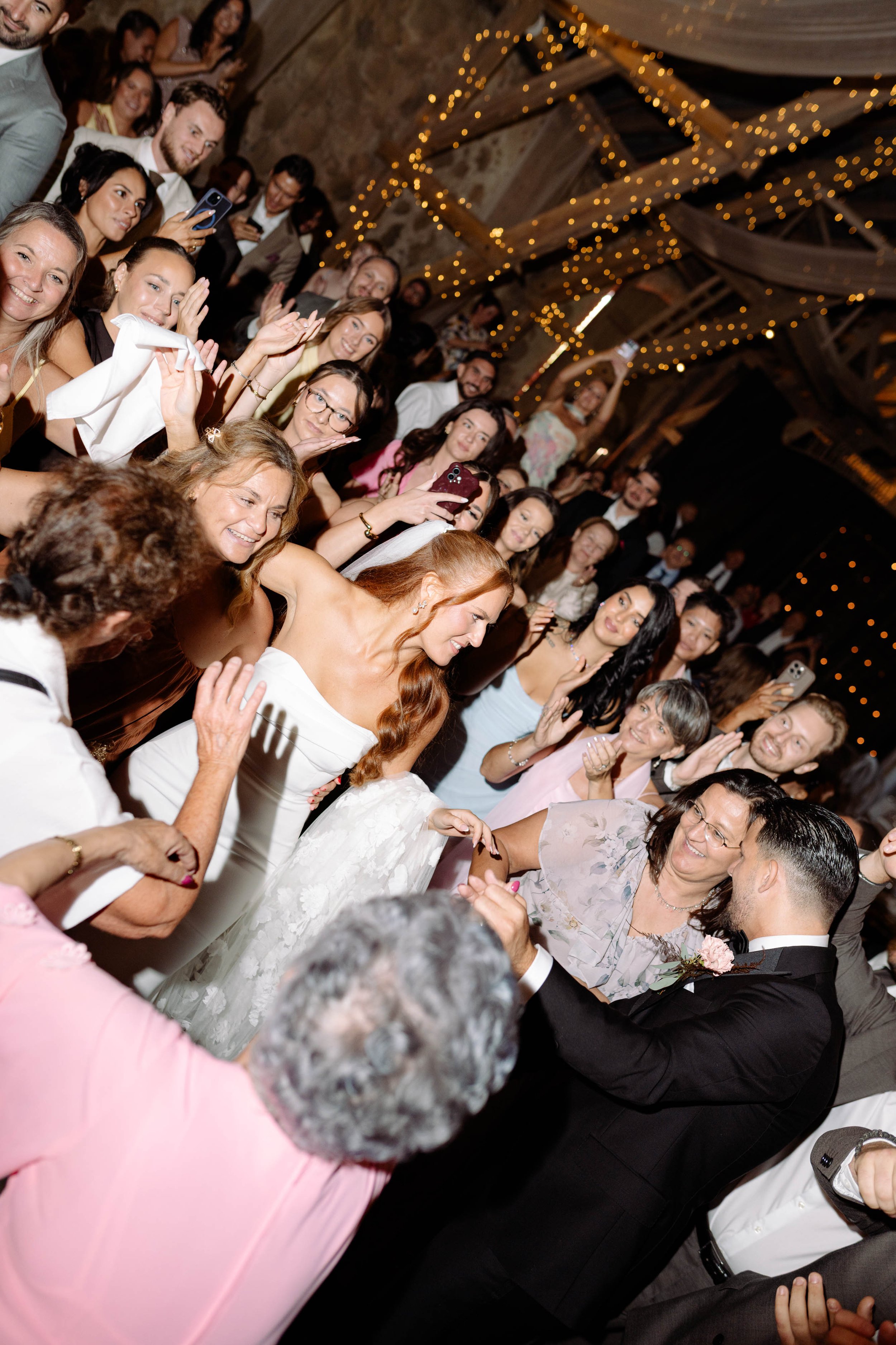 A bride and groom dancing at a wedding reception surrounded by family and friends in a rustic venue decorated with string lights.