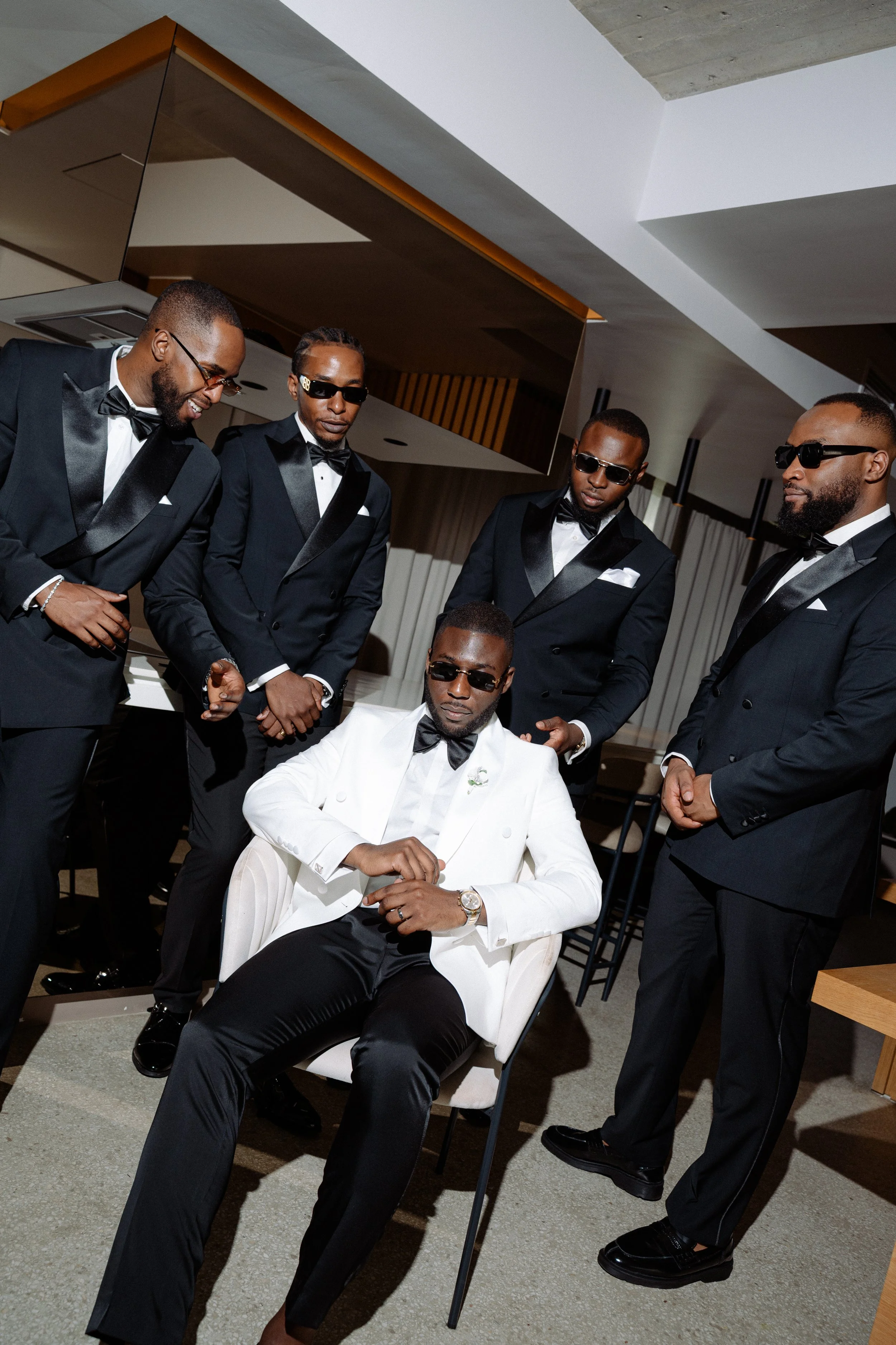 Six men dressed in formal black tuxedos and sunglasses, one seated in a white tuxedo jacket, the others standing around him, all inside a modern room.