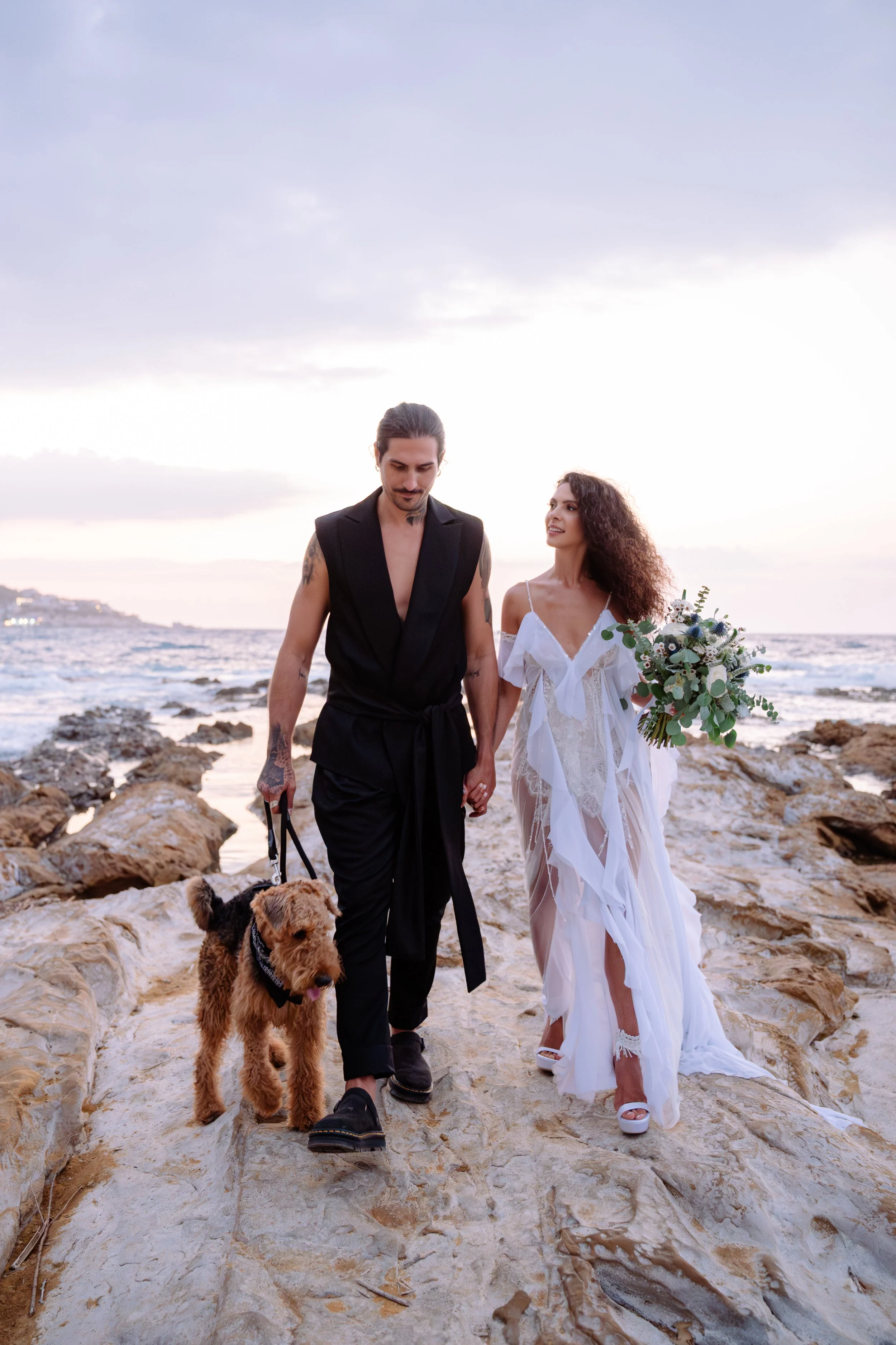 A couple dressed in wedding attire walking along a rocky beach at sunset, with a man holding a dog on a leash and a woman holding a bouquet of flowers.