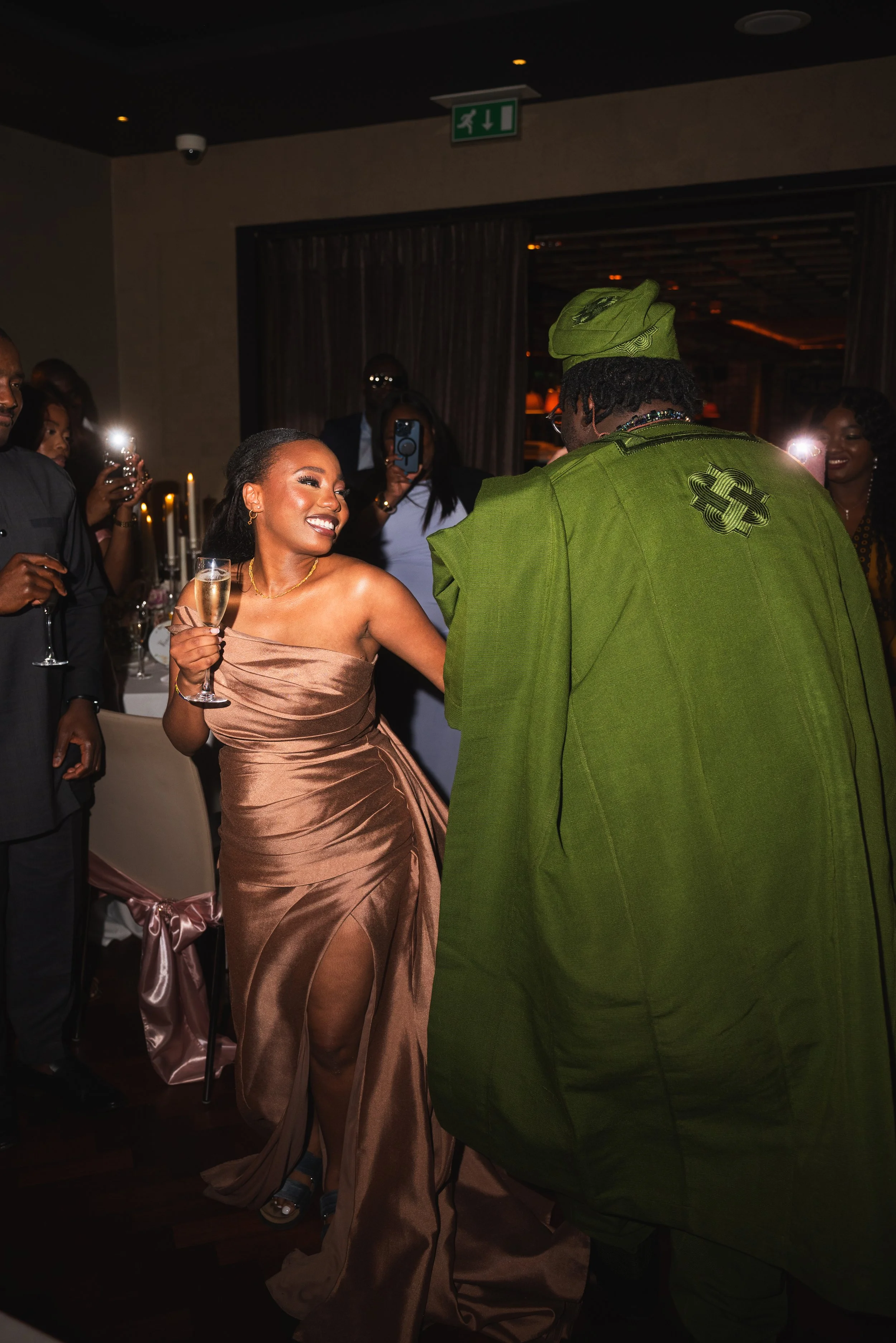 A woman in a strapless brown satin gown is smiling and holding a glass of champagne while dancing with a man dressed in traditional green attire at a celebration or party.