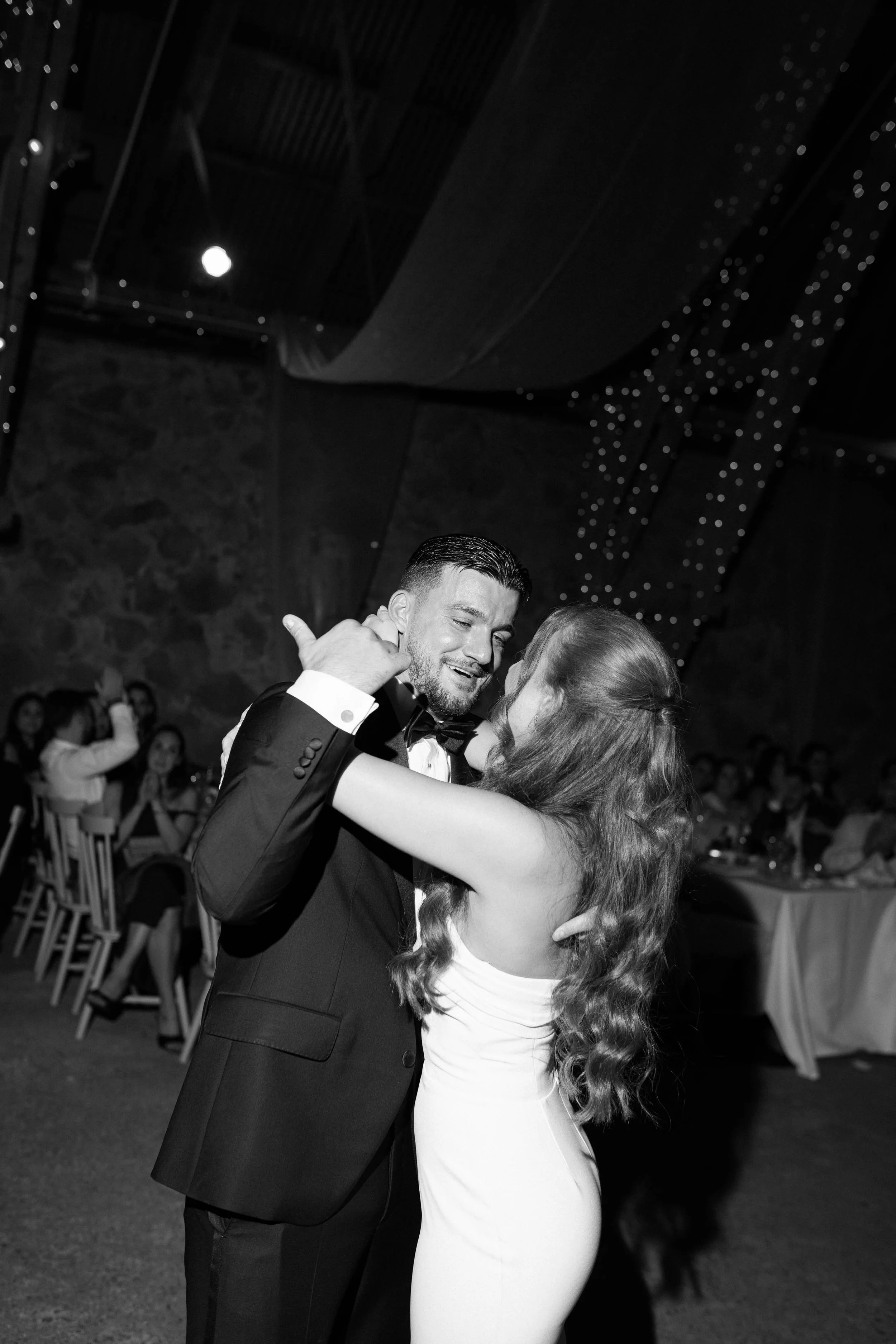 A couple dancing closely at a formal event, man in a tuxedo and woman in a strapless dress, with guests seated in the background.