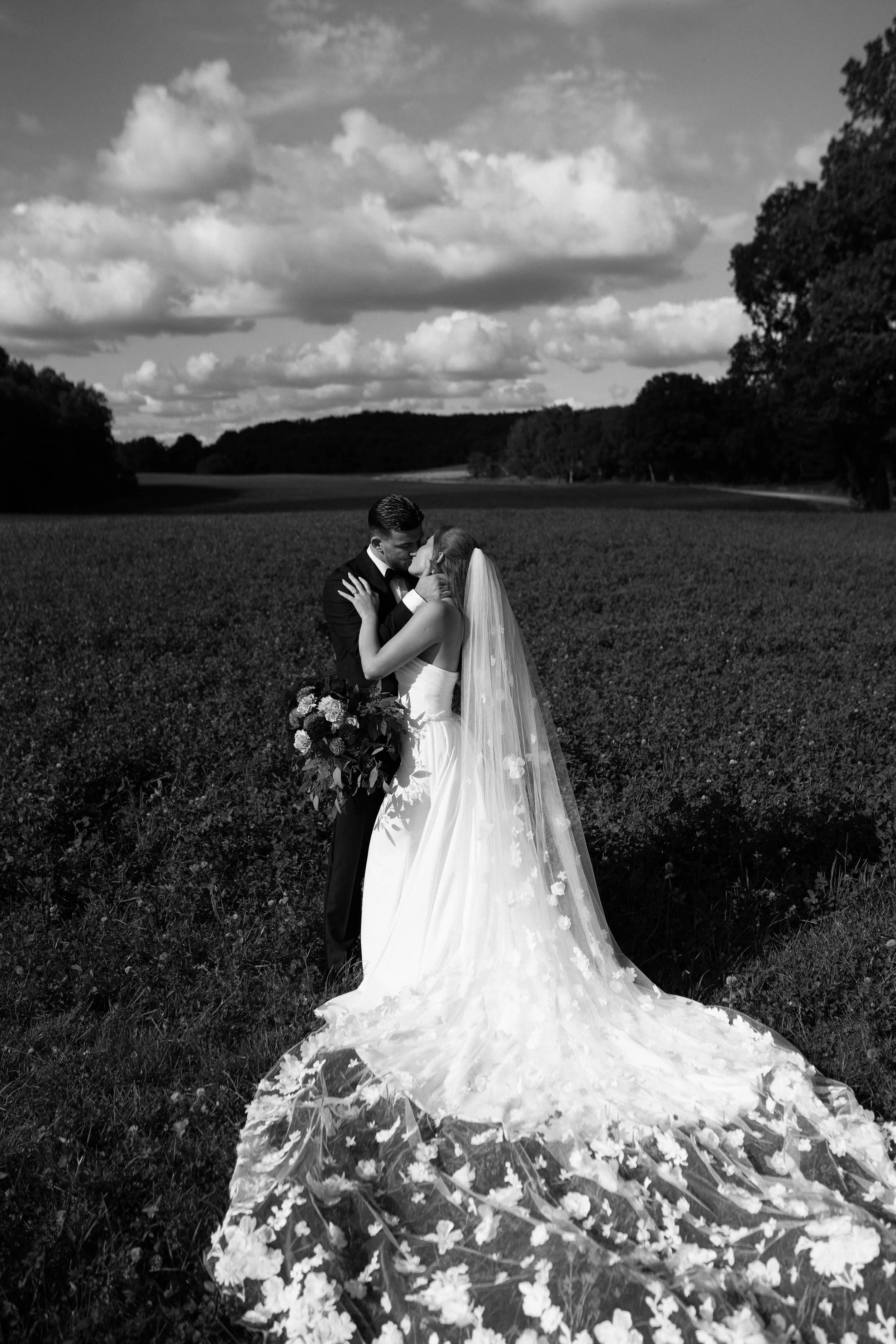 A black and white photo of a bride and groom kissing outdoors, with the bride holding a bouquet and wearing a long veil and dress, standing in a field with trees and a cloudy sky in the background.