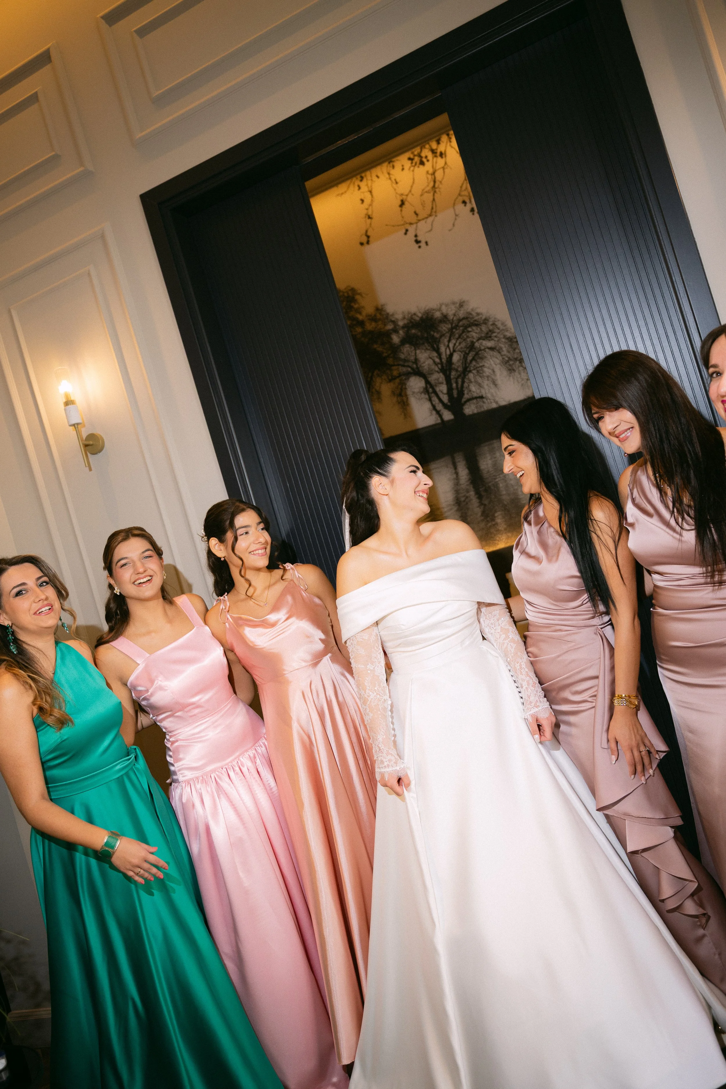 A bride and six bridesmaids smiling and standing together inside a decorated room with a window showing a tree and river view.