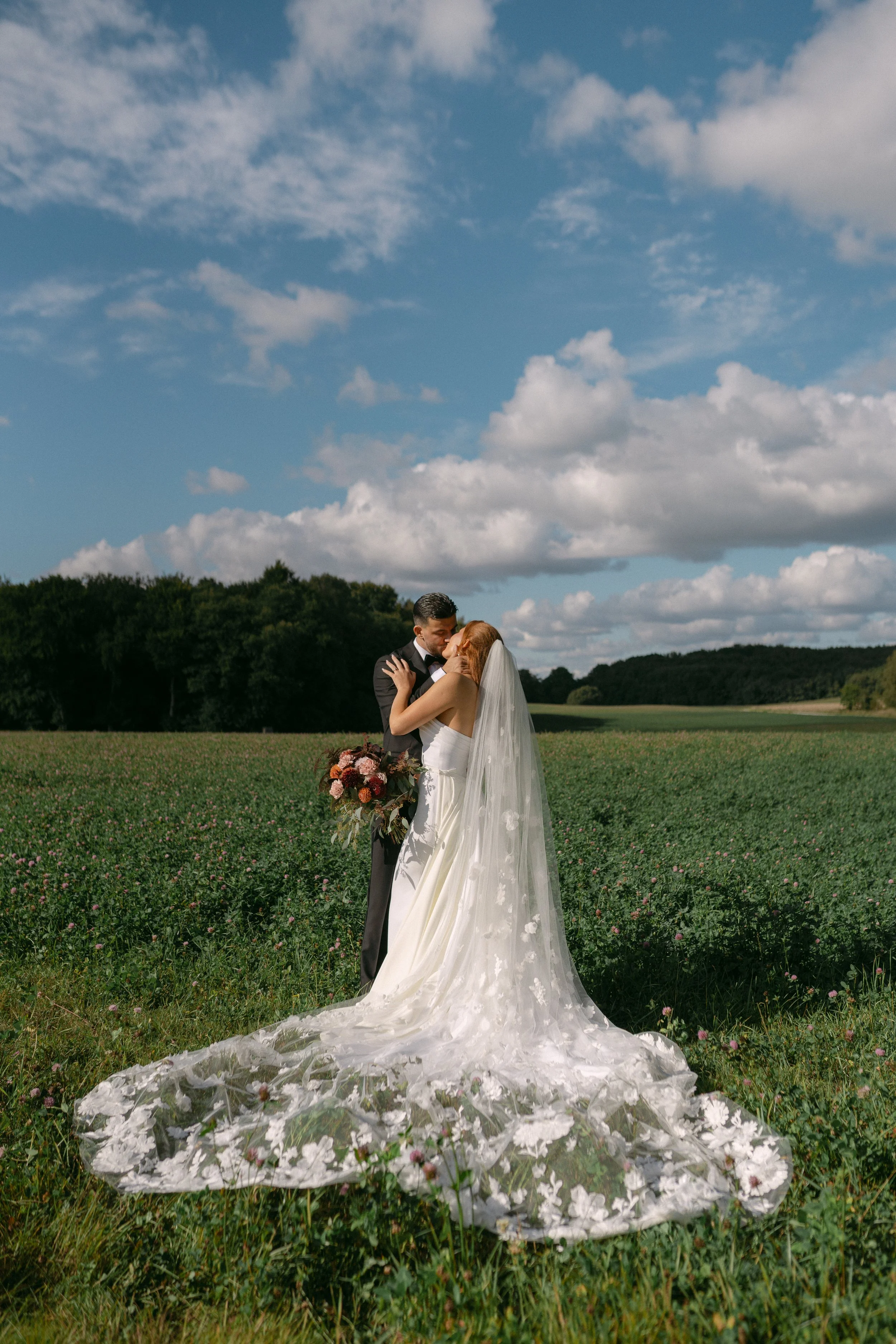 Bride and groom embracing in a field on their wedding day, the bride wearing a white gown with a long veil, holding a bouquet, the groom in a black suit, under a partly cloudy sky.