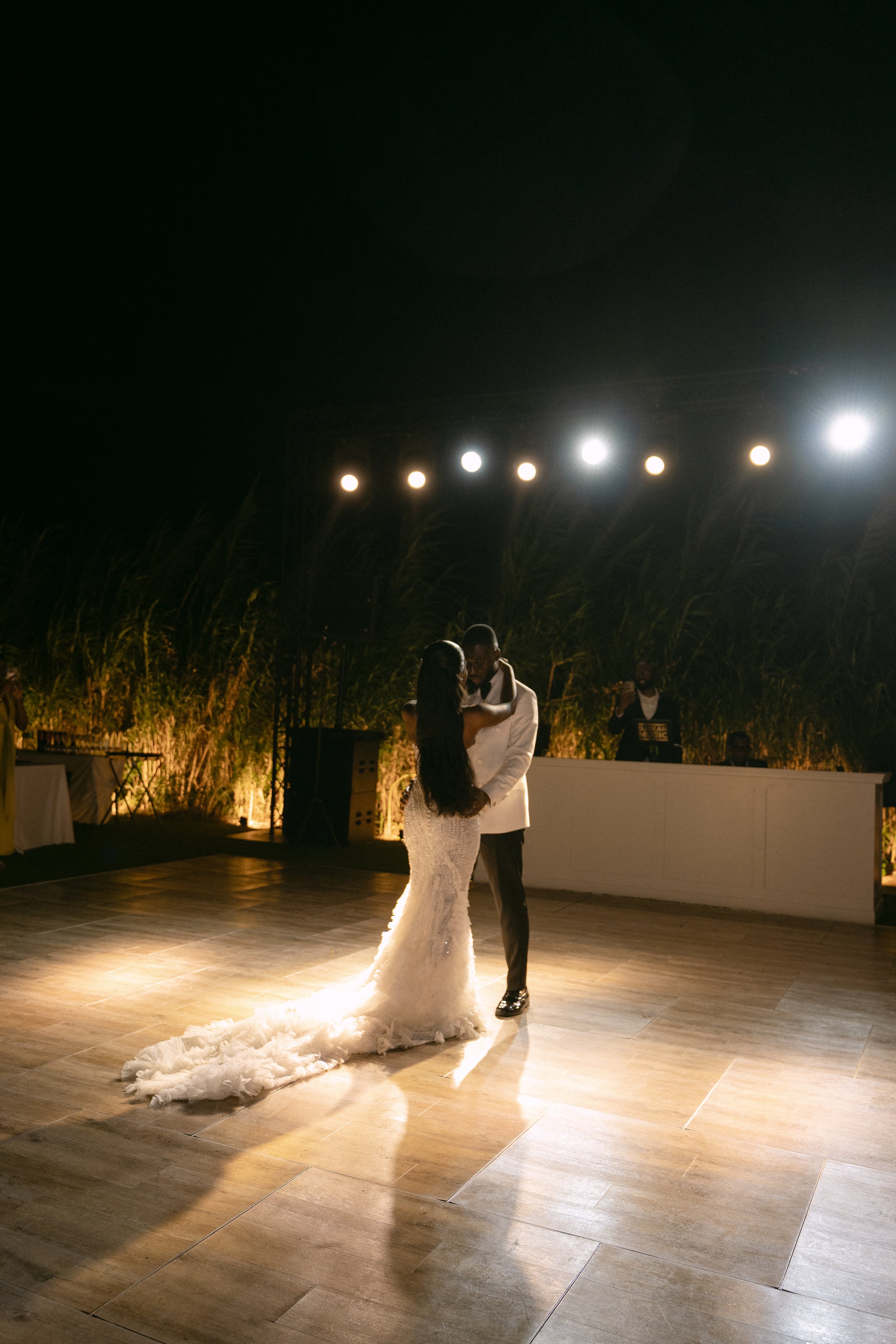 A bride and groom share a dance on a wedding dance floor at night, illuminated by bright overhead lights, with a DJ in the background.