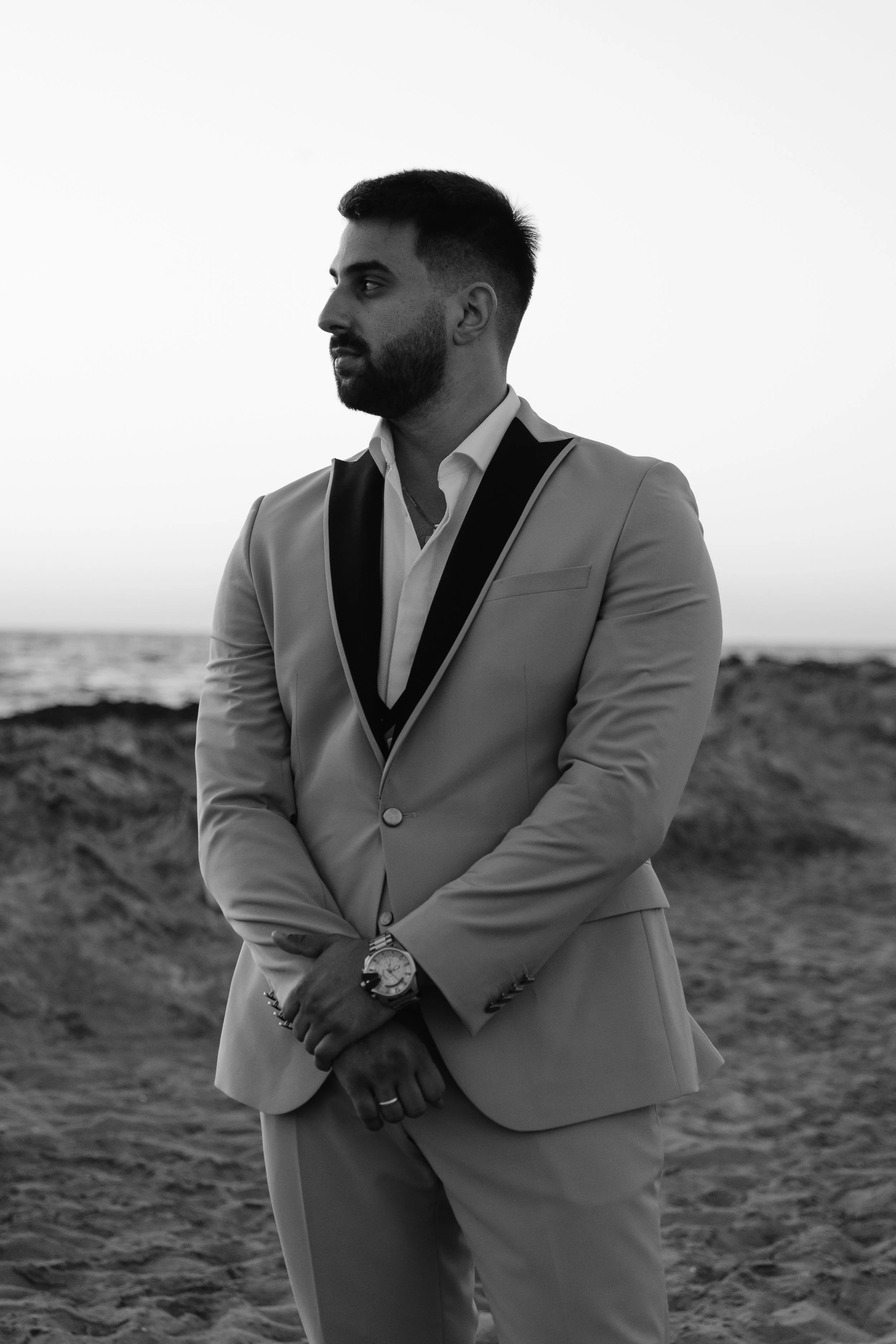 A man in a light-colored suit with a black lapel stands on the beach during sunset or sunrise, facing left with arms crossed, wearing a watch and rings, with sand dunes and the ocean in the background.