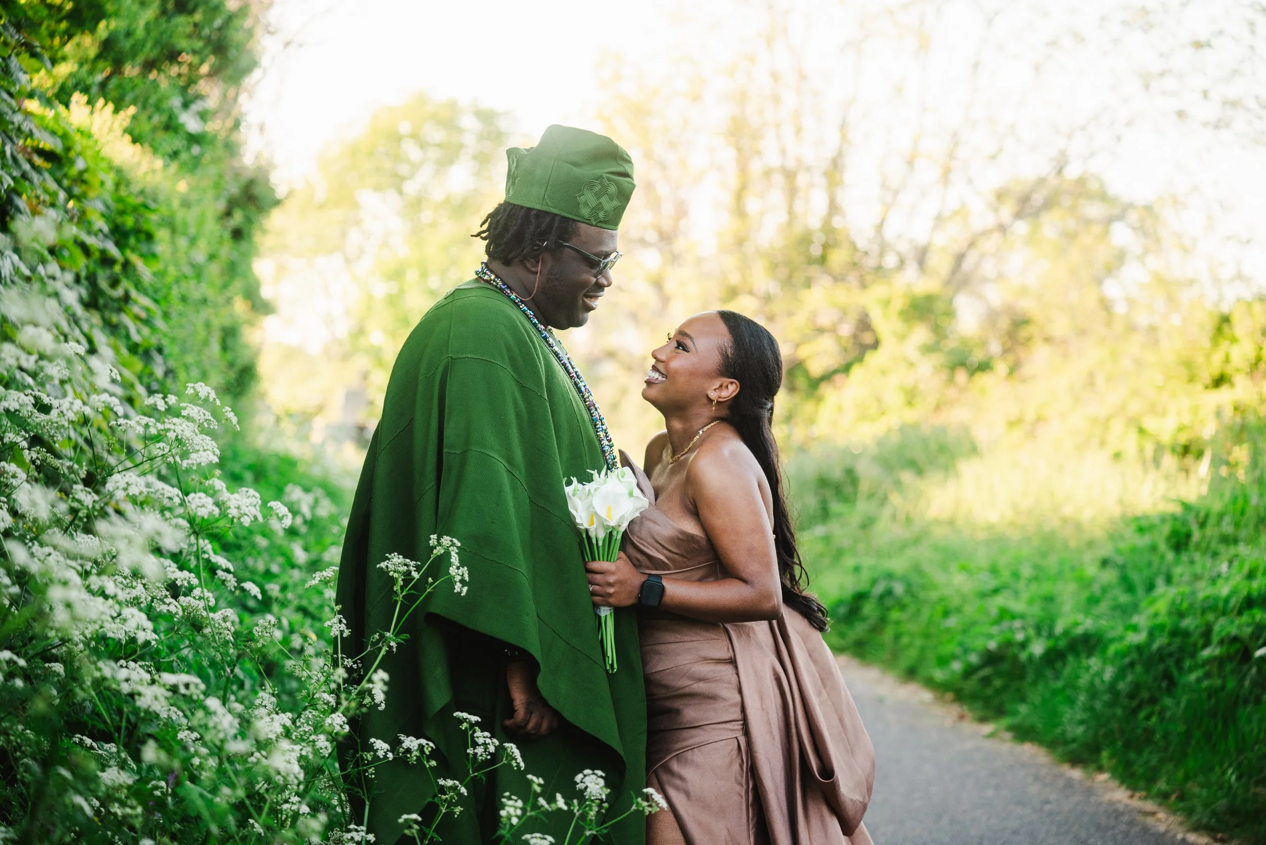 A couple stands close together outdoors on a peaceful path surrounded by greenery, with the man wearing a green robe and cap and holding a bouquet of white flowers, and the woman wearing a strapless brown dress, smiling at each other.