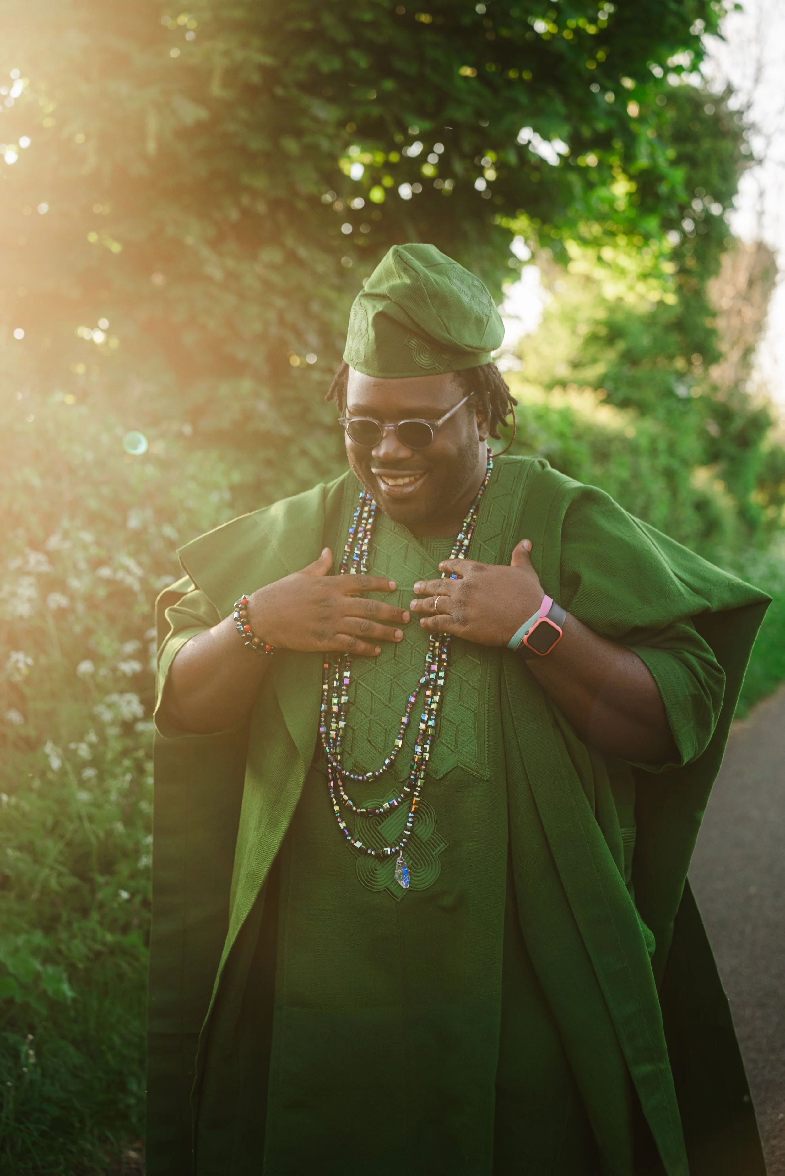 A person dressed in green traditional attire with beads and jewelry, wearing sunglasses and a hat, standing outdoors in sunlight surrounded by greenery.
