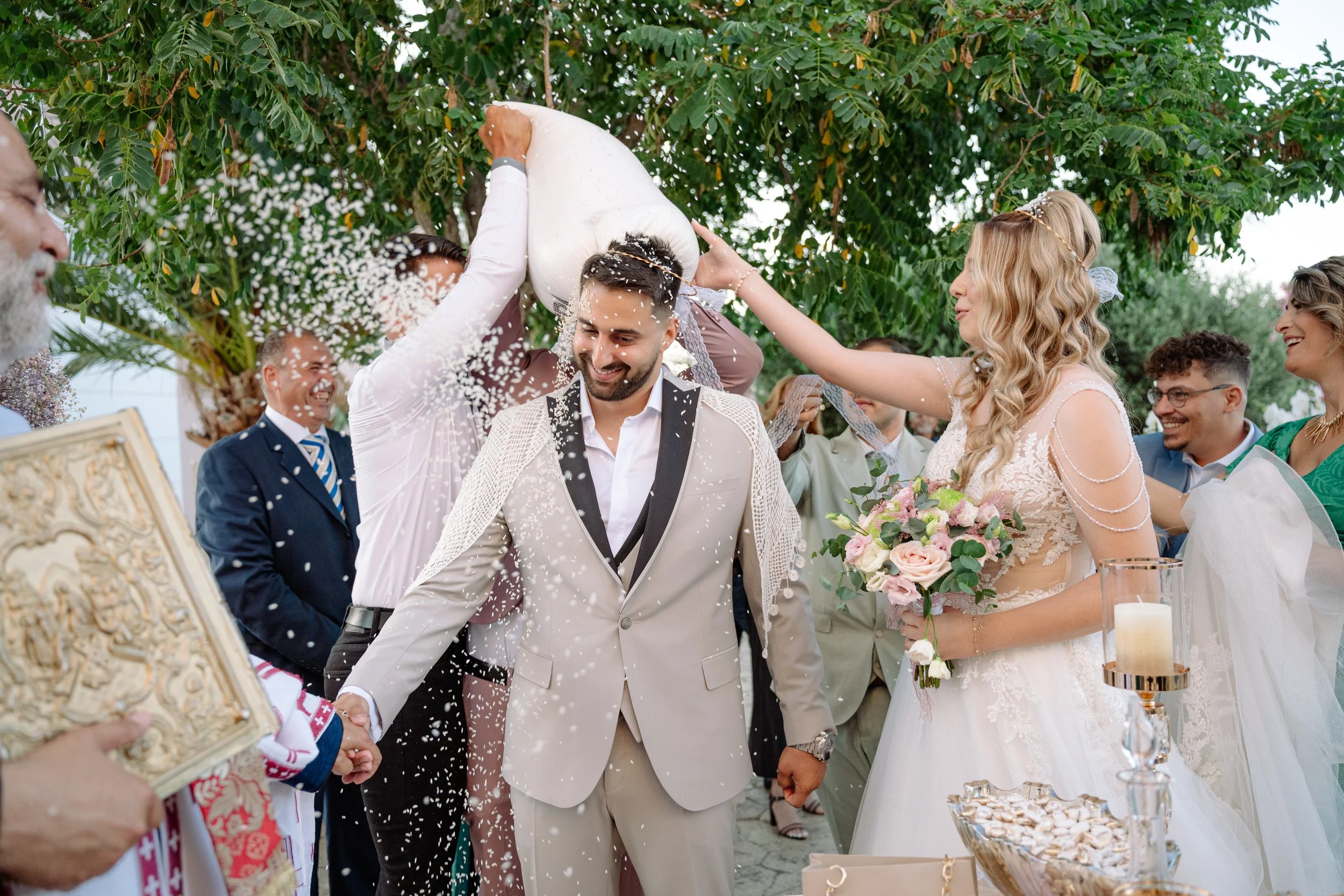 A wedding scene where the bride and groom are surrounded by friends and family. The groom is smiling as a person pours rice over his head, and the bride holds a bouquet while smiling. Guests are celebrating outdoors under a tree.
