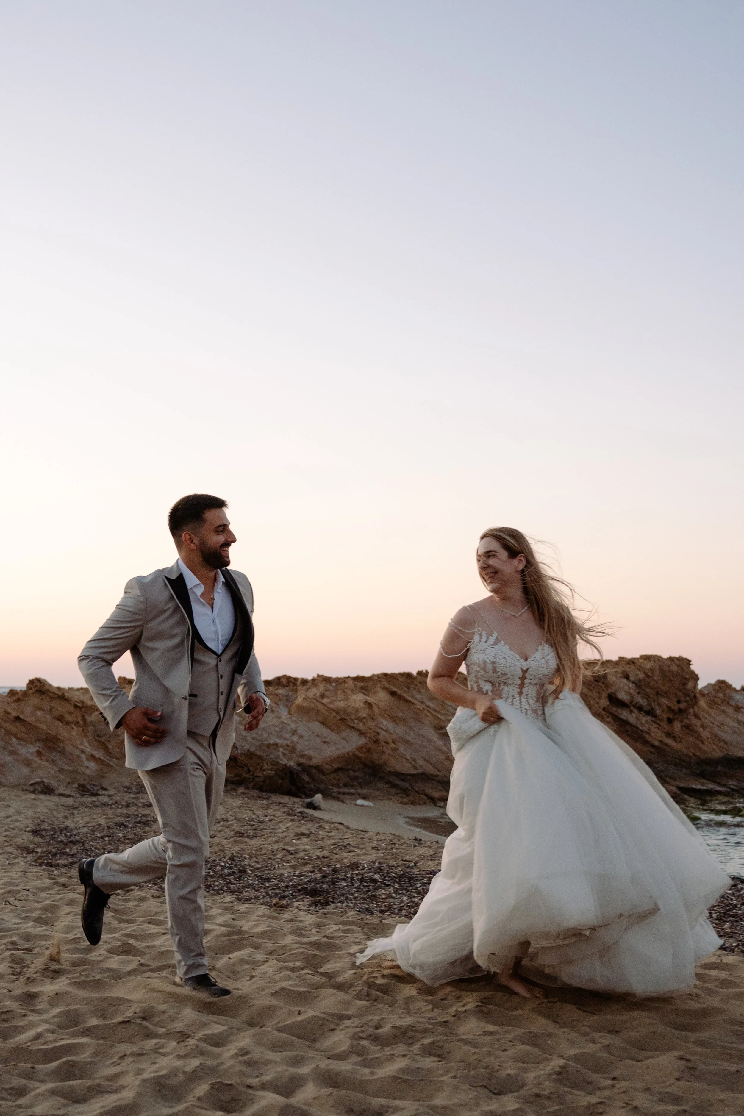 A newlywed couple running on a sandy beach at sunset with rocky cliffs in the background, the bride in a white wedding gown and the groom in a light gray suit.