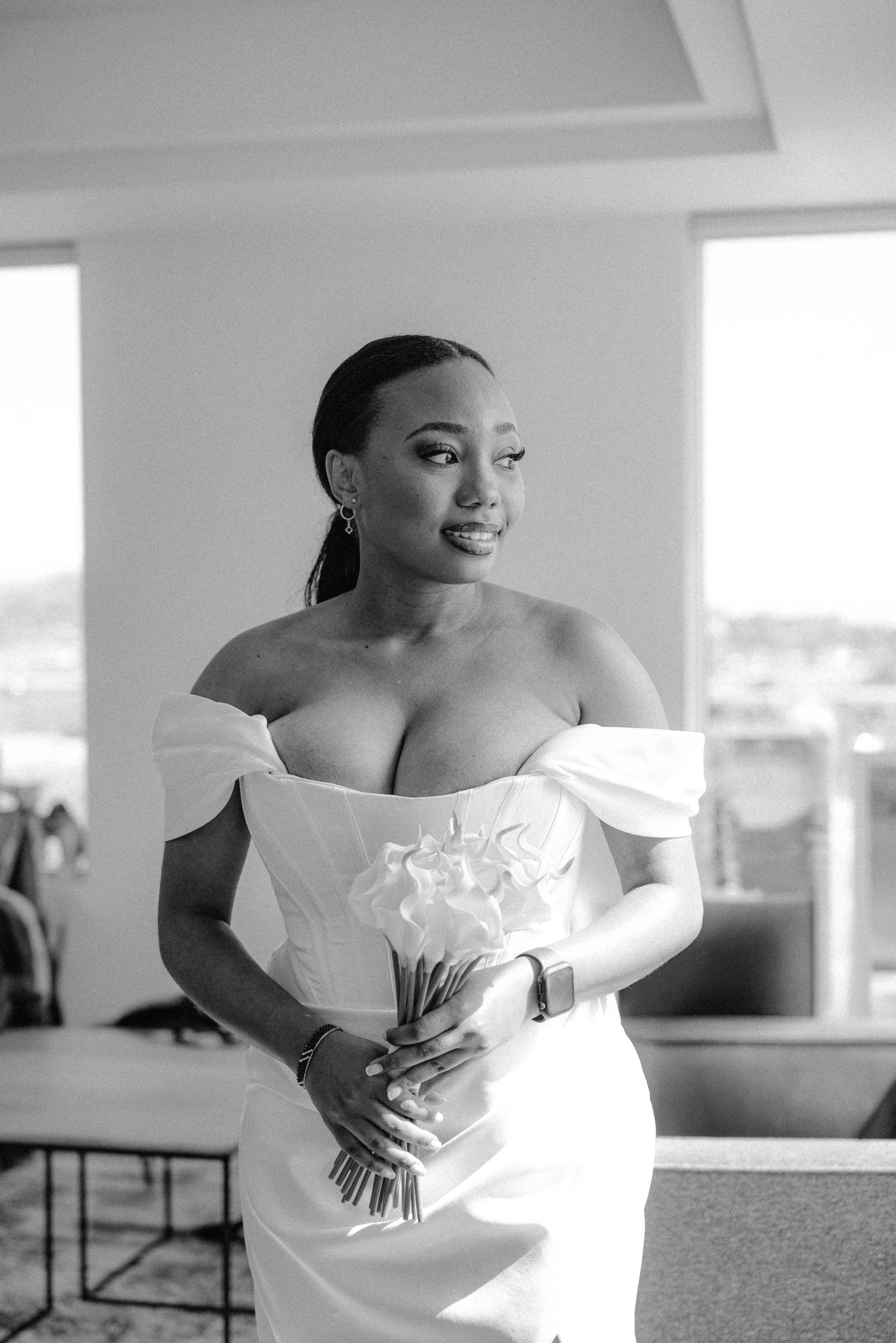 A woman in a wedding dress holding a bouquet of flowers, standing indoors with large windows in the background.
