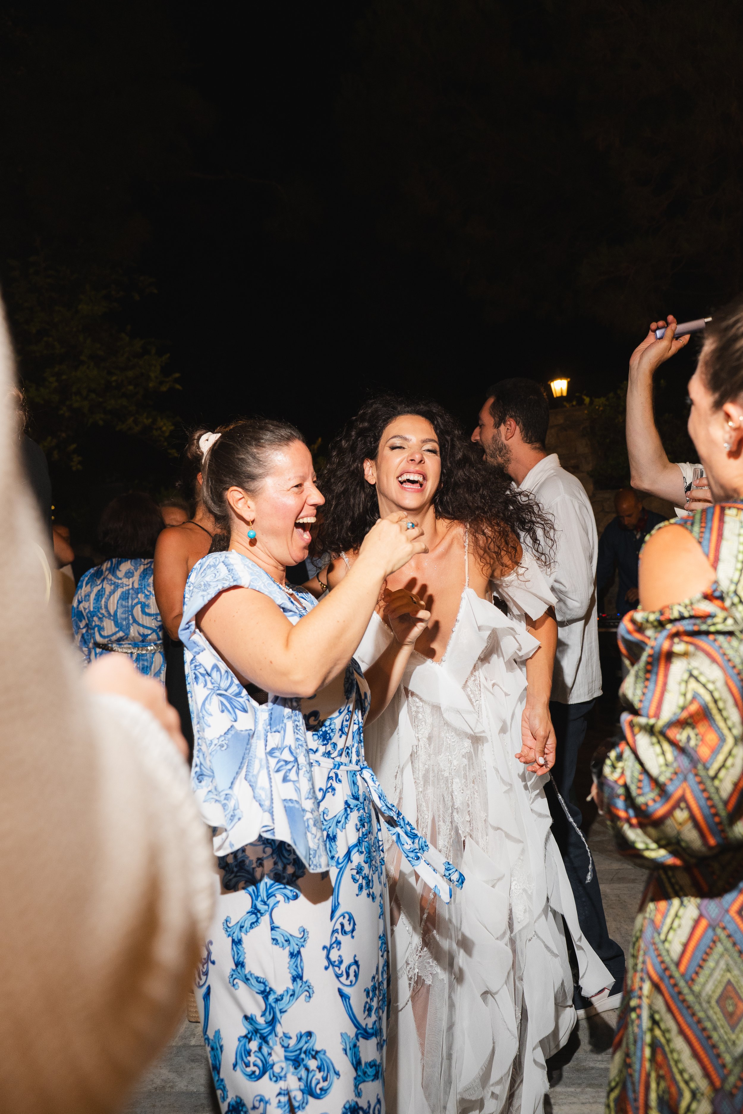 Group of people dancing and laughing at an outdoor party at night, with women wearing white and patterned dresses and appearing joyful.