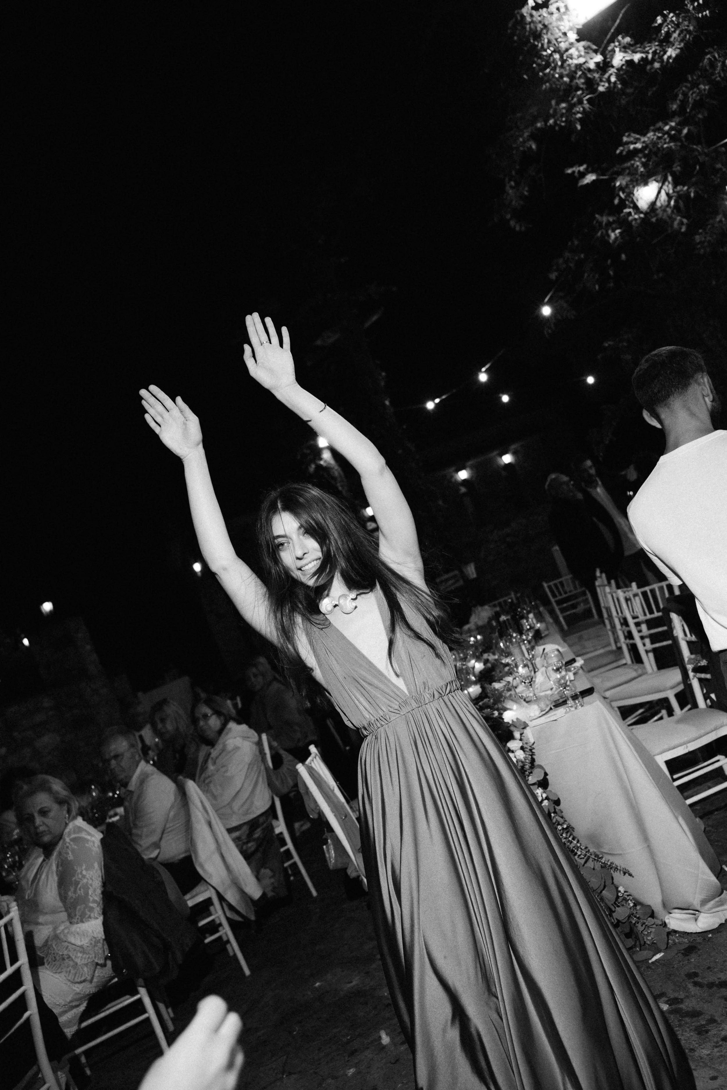 A woman in a long dress is dancing and smiling with her arms raised at a nighttime outdoor event, with guests seated at tables behind her.