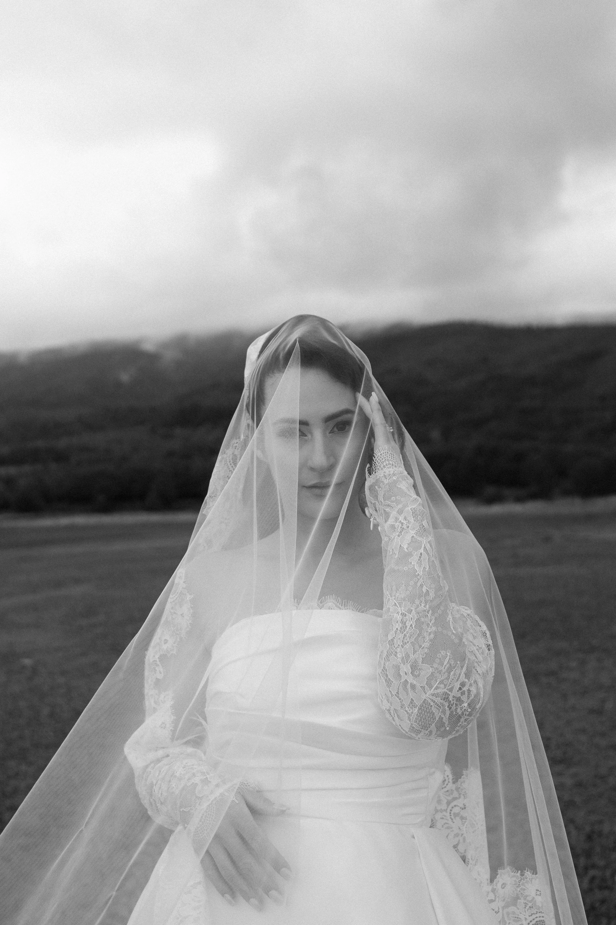 A woman in a wedding dress and veil standing outdoors with mountains in the background, holding her veil with one hand, in black and white.