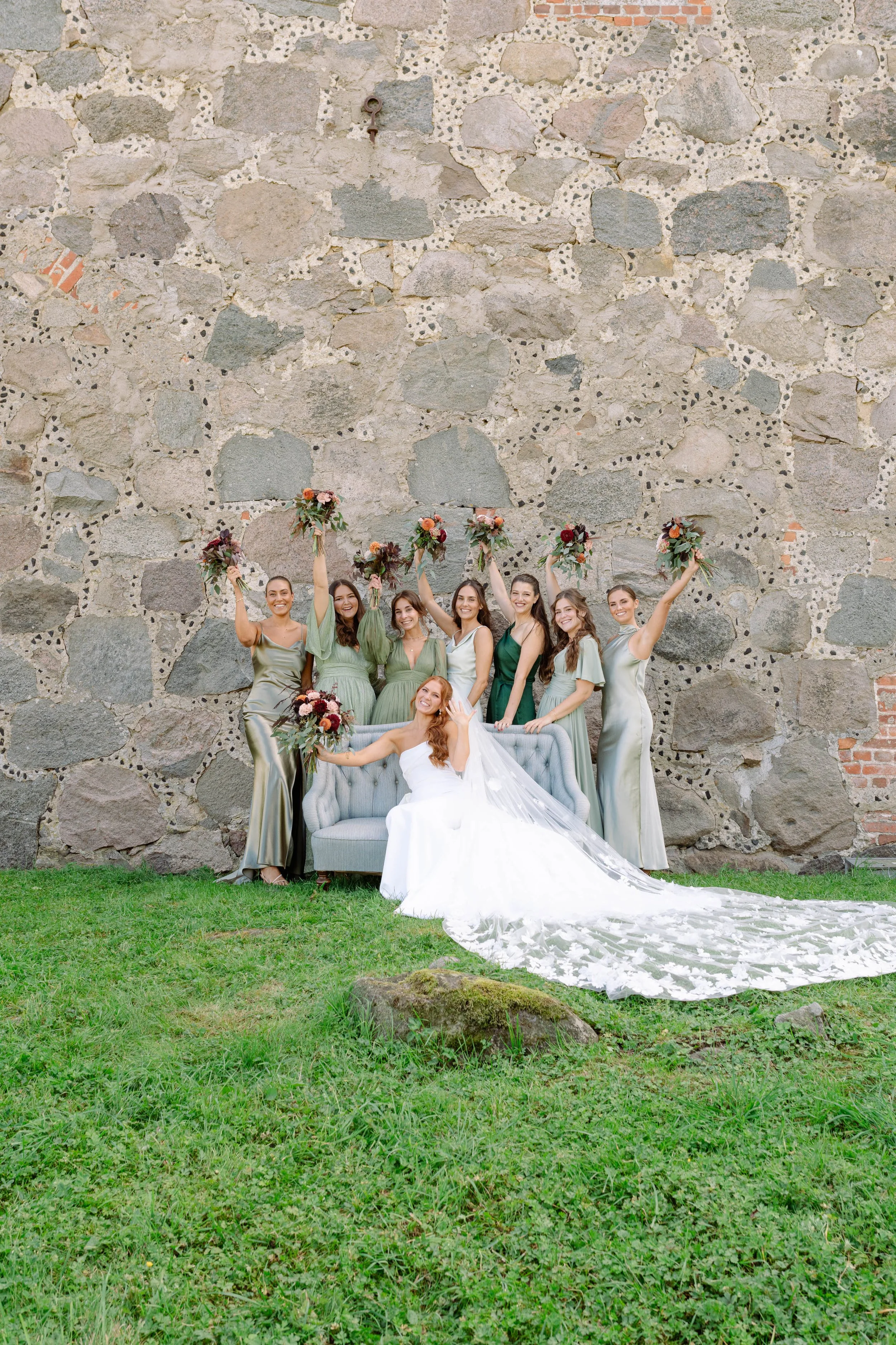A bride with long hair in a white wedding dress and veil sitting on a vintage sofa in front of a large stone wall, surrounded by eight women in green and beige dresses holding bouquets, celebrating at an outdoor wedding.