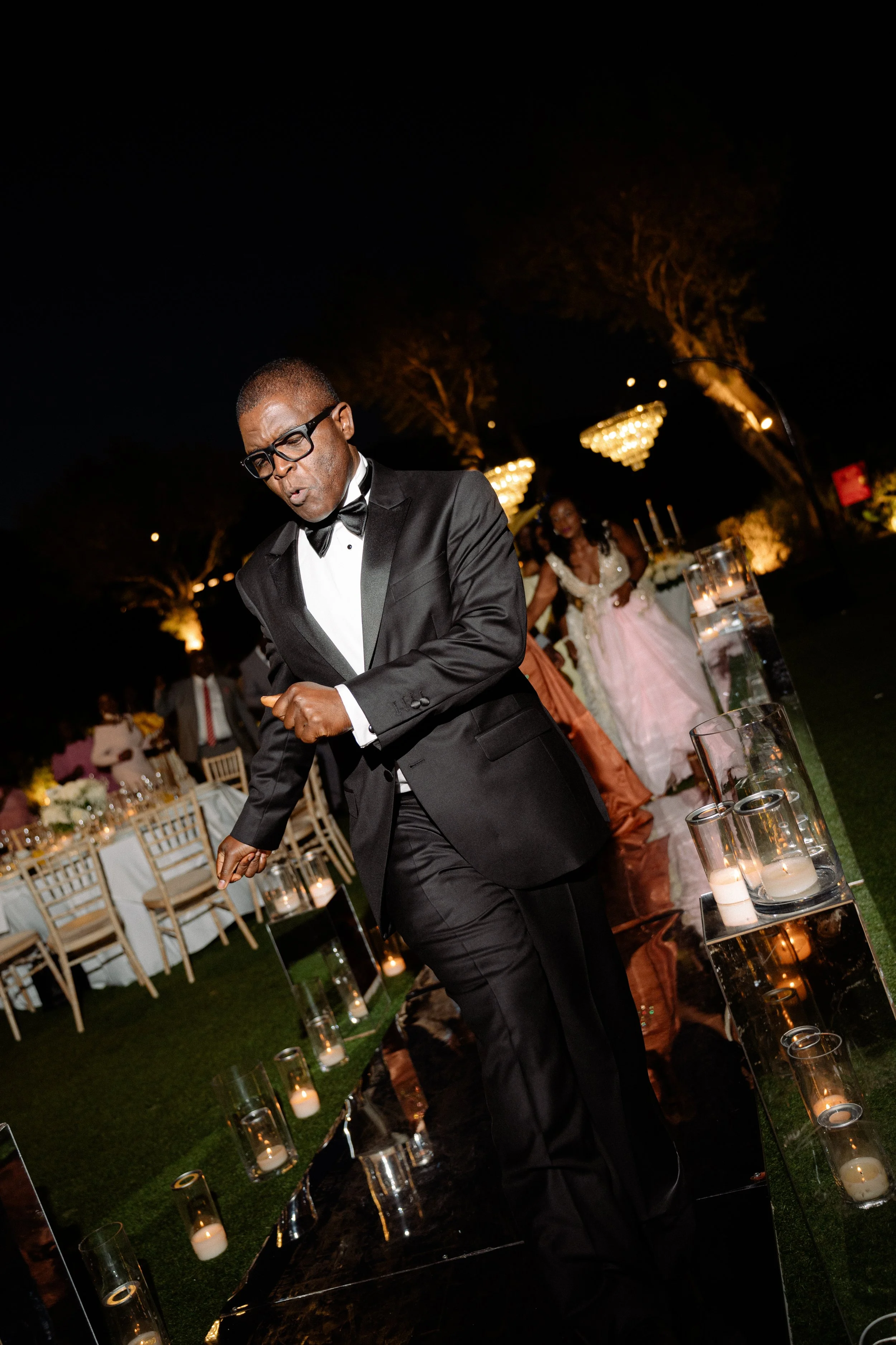 A man in a tuxedo dancing at an outdoor event during nighttime, with tables, chairs, and a woman in a pink dress in the background, decorated with candles and warm lighting.