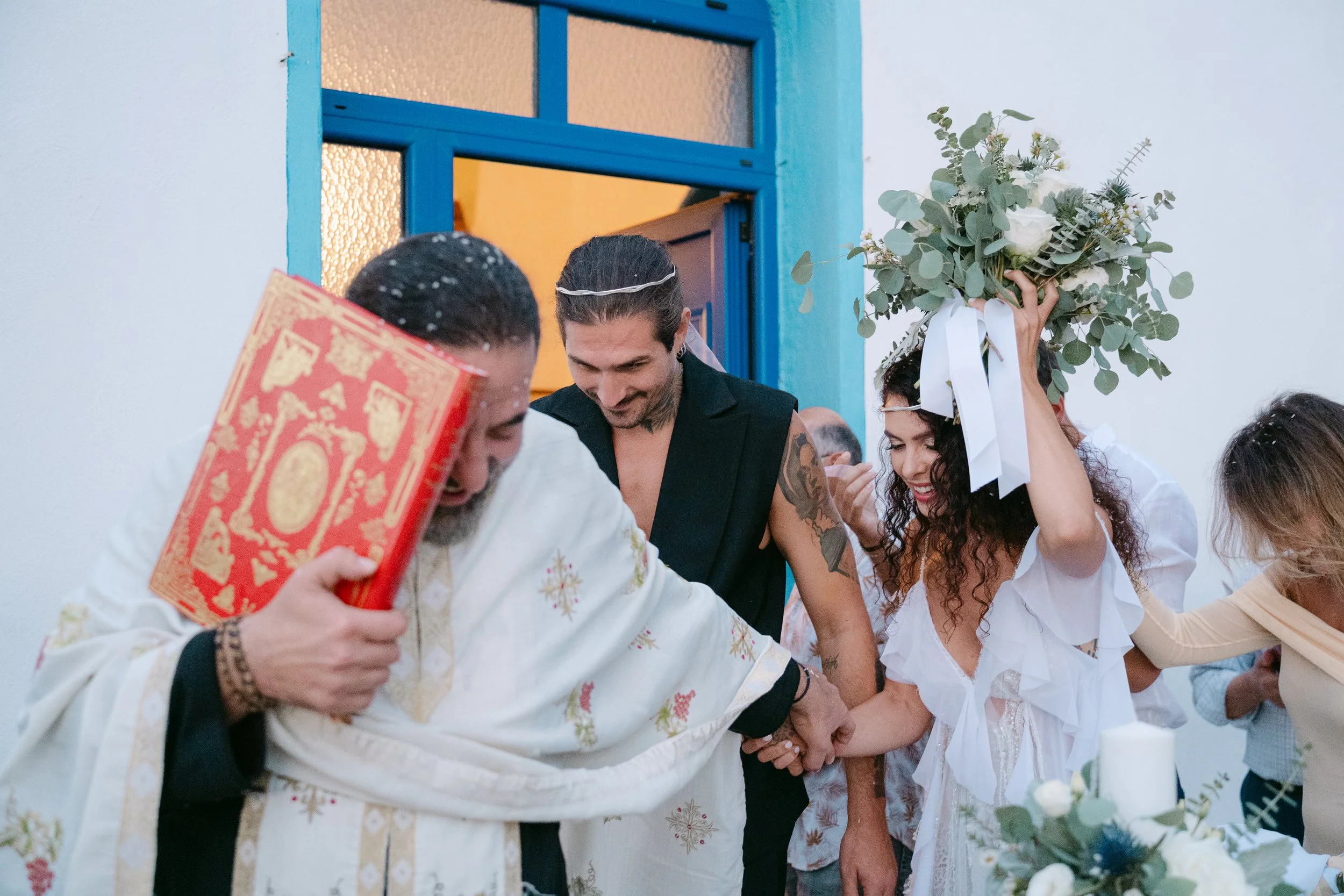 People celebrating a wedding inside a church, with the bride holding a bouquet of flowers and the groom wearing a sleeveless shirt, as the priest holds hands with them.