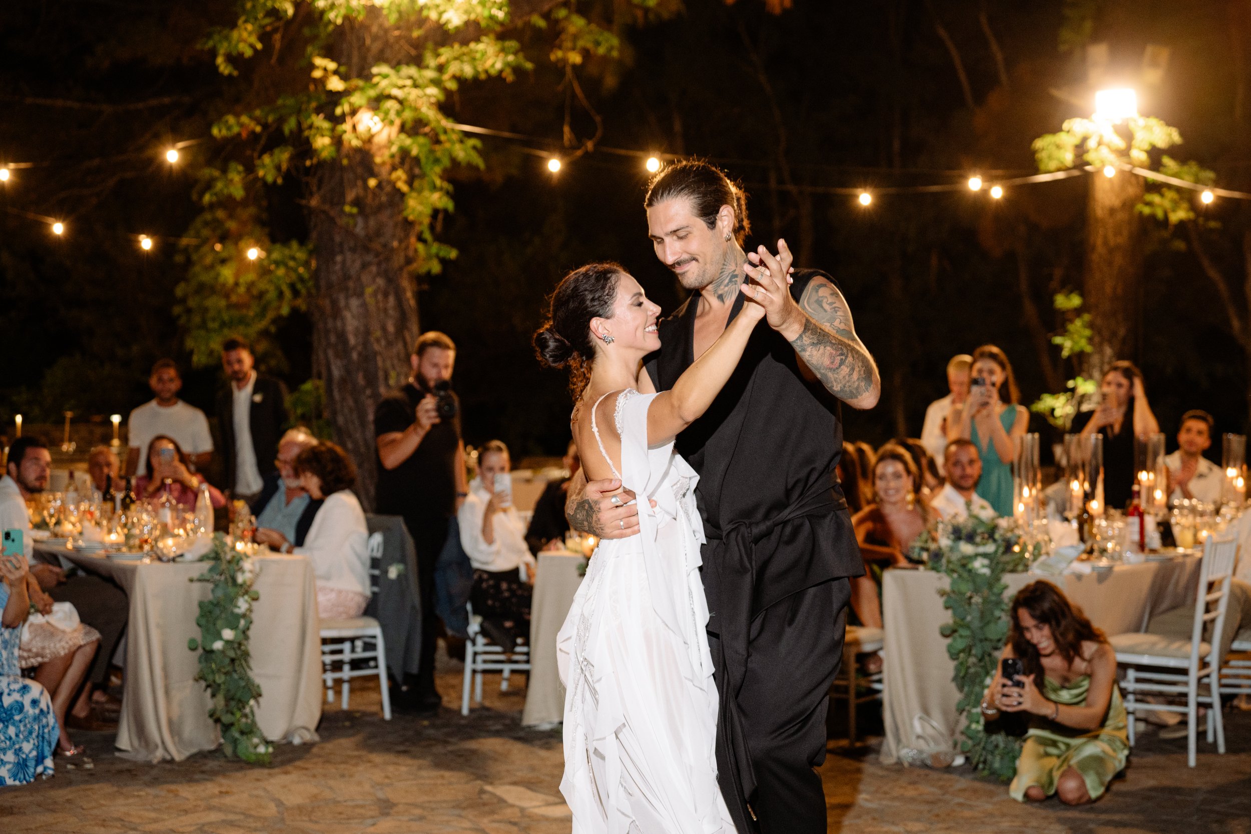 A bride and groom are dancing at their wedding reception outdoors at night. The bride wears a white dress, and the groom wears black. They are smiling and looking at each other. Guests seated at decorated tables and standing around are watching and t