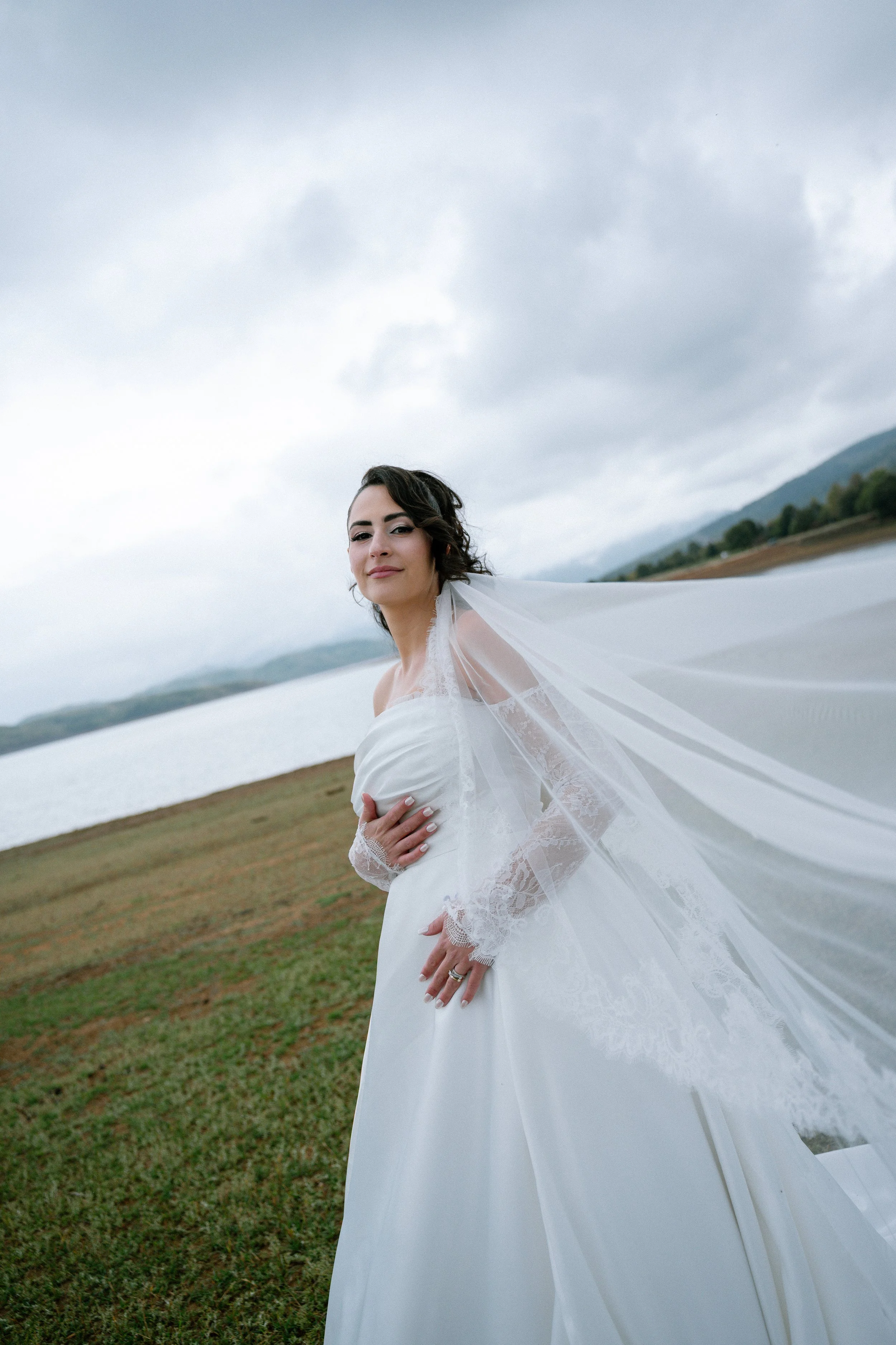 A bride wearing a white wedding dress and veil stands outdoors along a lakeshore, with mountains and a cloudy sky in the background.