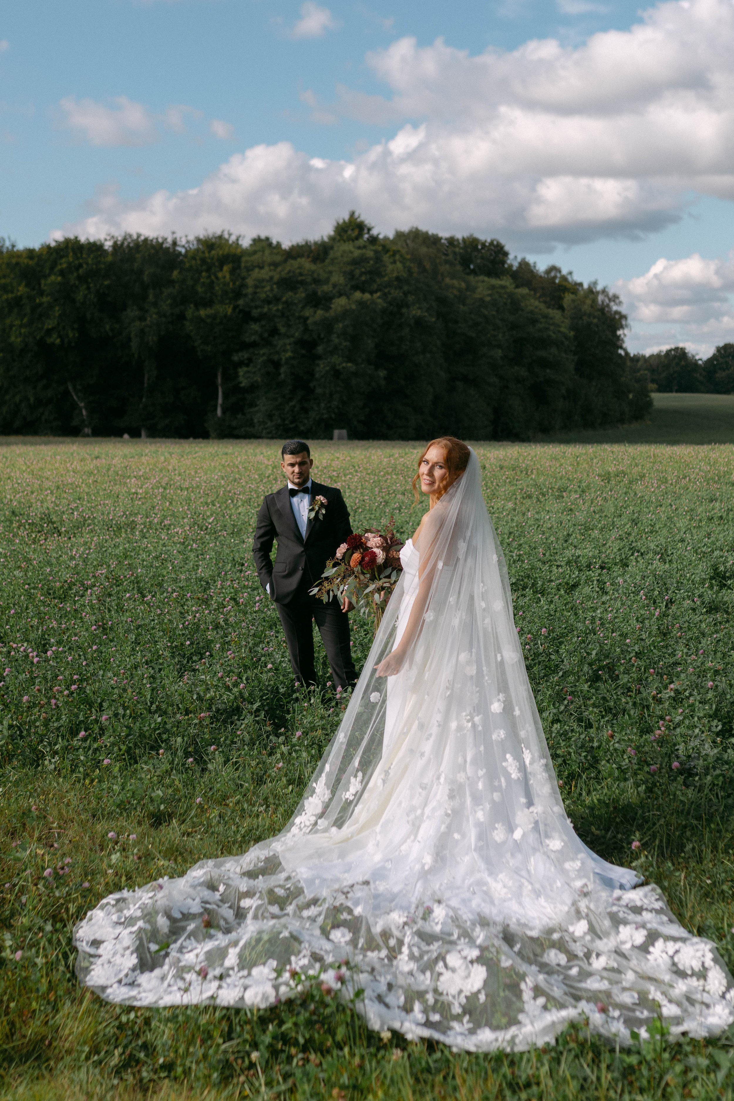 A bride and groom stand in a field on their wedding day. The bride wears a long, flowing white wedding gown with a veil, holding a bouquet of flowers. The groom is dressed in a black tuxedo with a bow tie, holding a small bouquet or similar floral ar