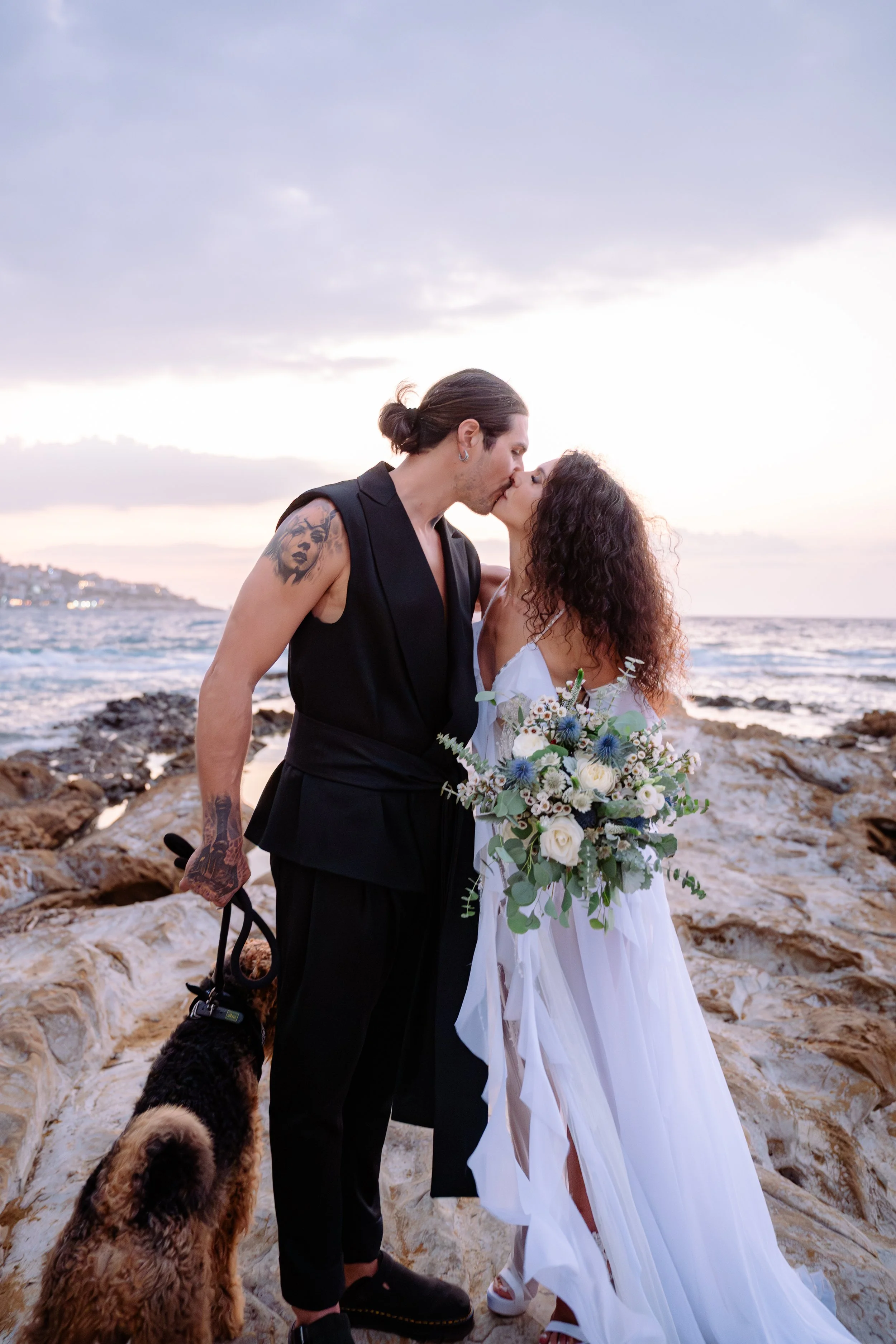 A couple dressed in wedding attire sharing a kiss on a rocky beach at sunset, with a bouquet of white and blue flowers, and a dog beside them.