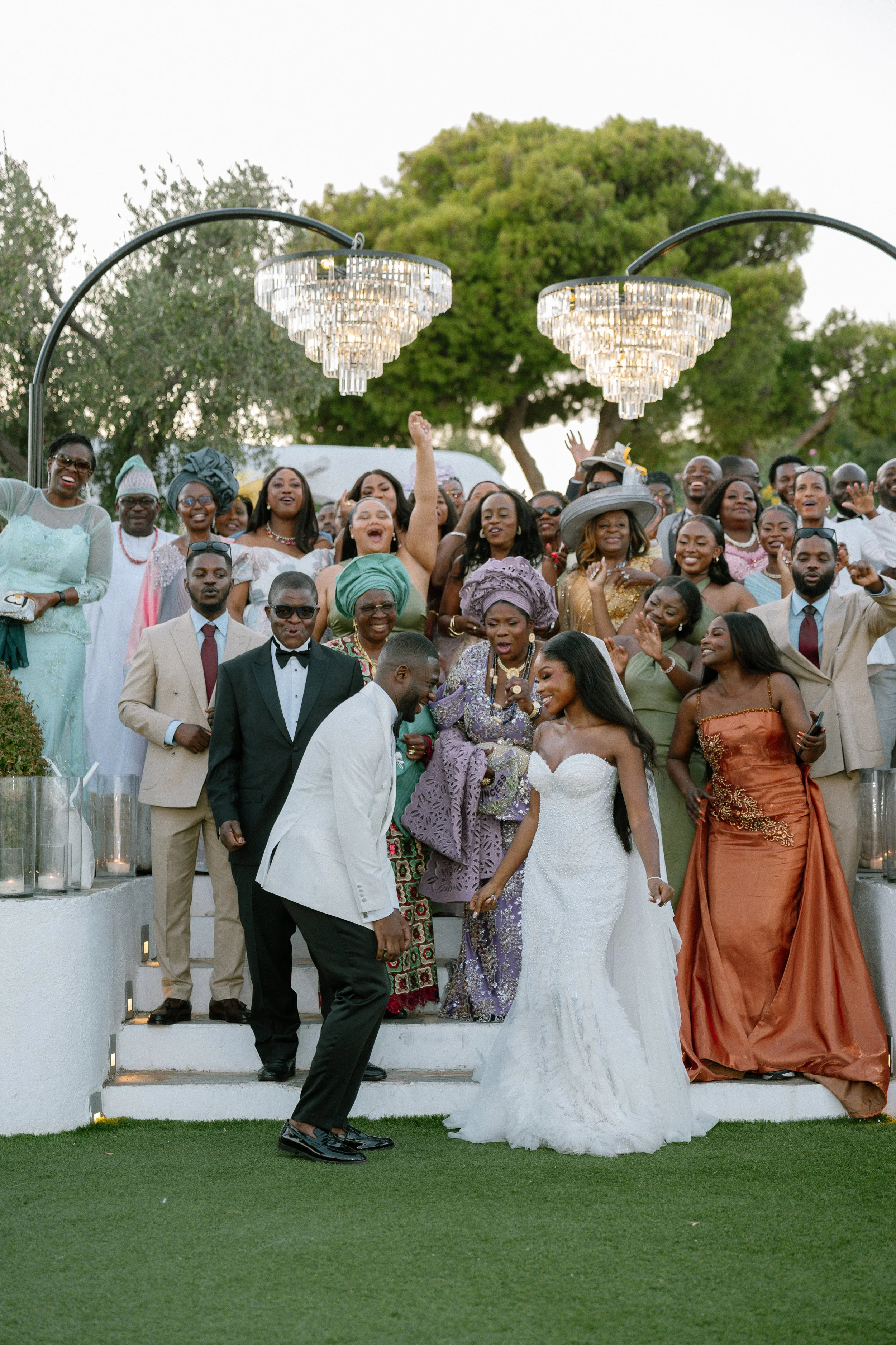 A large group of people dressed in formal and traditional attire at an outdoor wedding celebration, with a bride and groom in the foreground and nature in the background.